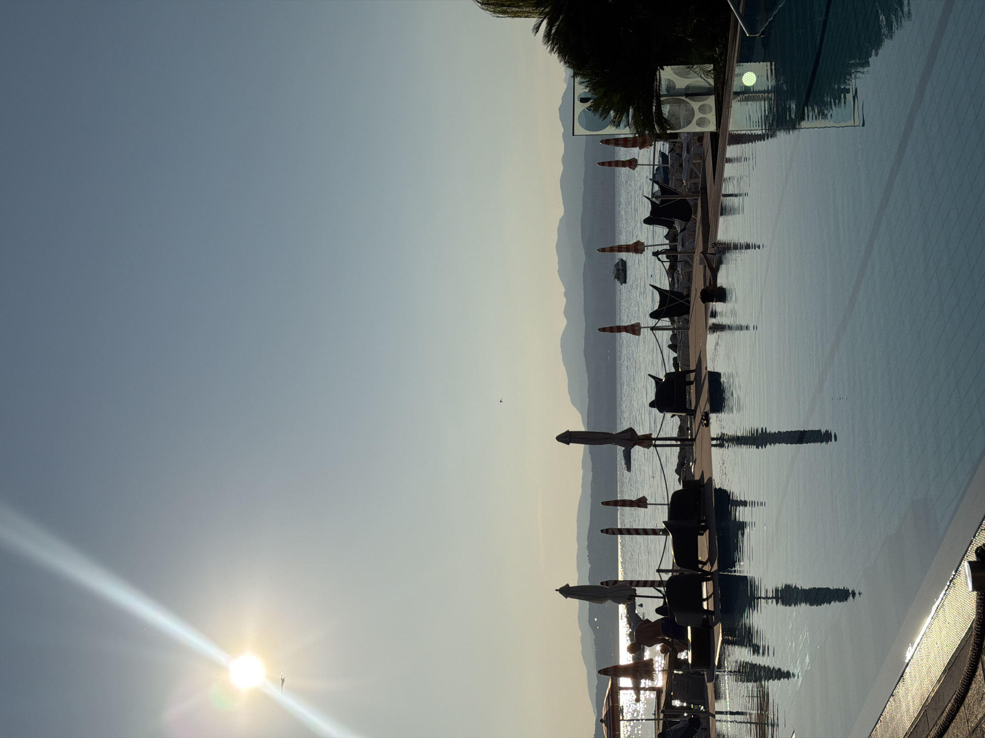 This is a vertical, eye-level, medium shot of a swimming pool area on a sunny day, with a lake and distant mountains in the background. The sun, positioned in the upper left quadrant, is very bright, casting a lens flare and a visible ray of light. A small airplane is visible in the sky near the sun. The swimming pool occupies the bottom half of the image, with its water reflecting the surrounding objects and the sky. The edge of the pool, a textured rope, and a metal bollard are visible in the foreground. Beyond the pool, a paved area is lined with lounge chairs and several closed umbrellas, some with red and white stripes. A few people are scattered around the lounge area and in the pool. In the distance, a calm lake stretches out, with boats on the water and a hazy mountain range forming the horizon. The overall scene suggests a resort or hotel area in Sirmione, Italy, likely during the daytime.