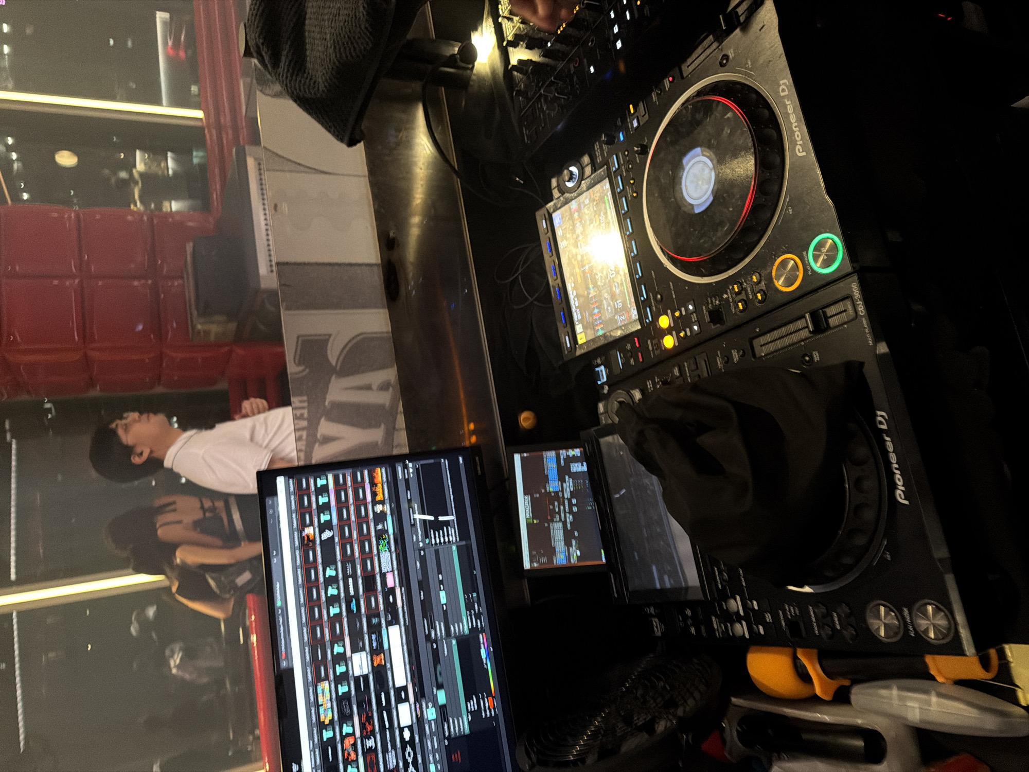 An indoor scene features a DJ setup in the foreground, with a man in a white collared shirt standing to the right, looking to the side. Behind him, two women are visible in silhouette, one wearing a black top with a bra strap visible. The background shows a large window overlooking a cityscape, with red stacked structures and illuminated lights. The DJ equipment includes a monitor displaying waveforms, a smaller screen, two CDJs, and a mixer. A black cloth is draped over part of the setup. The scene appears to be a nightclub or performance venue.