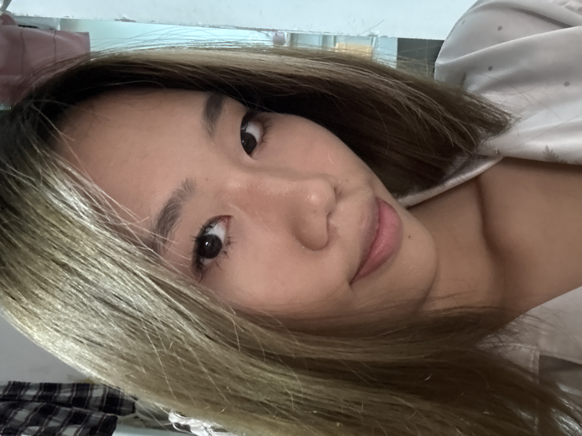 The primary subject of the image is a young woman of East Asian descent, captured in a close-up, angled portrait, likely a selfie. She is positioned with her head tilted to her right, gazing directly into the camera with a neutral expression and a subtle smile.
Her hair is long and flows around her face and shoulders. It is dyed a light brown or ash blonde, contrasting with darker roots visible near her scalp. Her facial features are distinct, with clear skin, dark eyes, defined eyebrows, and prominent eyelashes. Her lips are a soft pink, slightly parted.
She is wearing a light-colored garment, possibly a shirt or top, with a faint dotted pattern visible on the fabric. In the lower-left quadrant of the image, partially obscured by her hair, a piece of fabric with a black and white/grey plaid pattern can be seen, which might be another item of clothing or bedding.
The background is largely out of focus, indicating an indoor setting. Behind her, a light-colored surface, possibly a wall or headboard, is visible, alongside hints of a darker, perhaps pink, object in the upper left area. The overall lighting is soft and even, consistent with an indoor environment. No text is visible within the image.