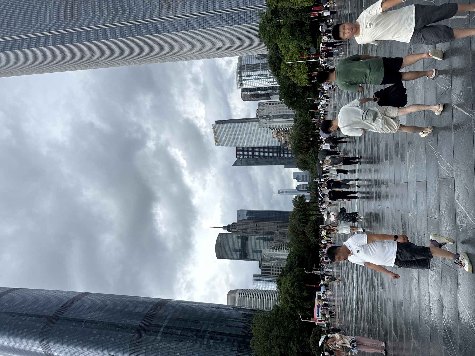 In Tianhe District, China, on a cloudy day, a wide, wet, tiled plaza is occupied by a crowd of people walking and standing. Two towering, modern skyscrapers flank the scene on the left and right, their reflective glass facades obscuring the overcast sky. In the mid-ground, a dense cluster of various other modern office buildings rises behind a line of lush green trees, including palm trees.
In the foreground, a man wearing a white polo shirt and dark shorts walks away from the camera, his legs slightly apart in a mid-stride pose. To his right, two young men walk together, one in a white t-shirt and shorts, the other in a green t-shirt and dark shorts. A young boy in a white t-shirt and grey shorts walks towards the camera on the far right. Many other individuals, some holding umbrellas, are dispersed throughout the plaza, indicating a public gathering or thoroughfare. The wet surface of the plaza suggests recent rainfall.
