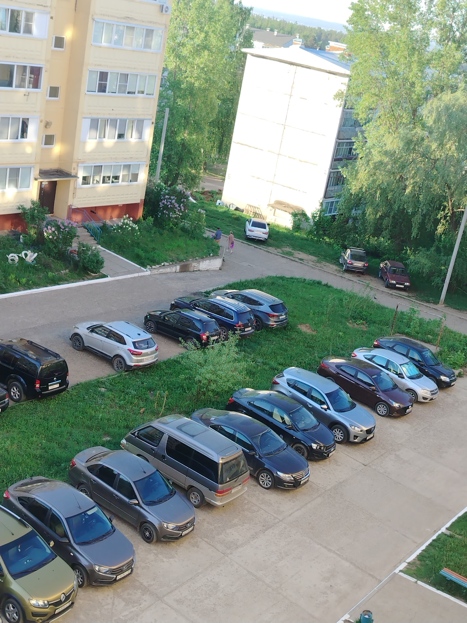 The image depicts an elevated view of an urban residential area in Noviy, Russia. Two multi-story apartment buildings with a pale yellow and white exterior are visible, surrounded by greenery. A concrete parking area contains numerous parked cars of various makes and models. A grassy area with trees separates the parking area from the road. In the distance, two people are walking along the road. The scene appears to be captured during daylight hours.