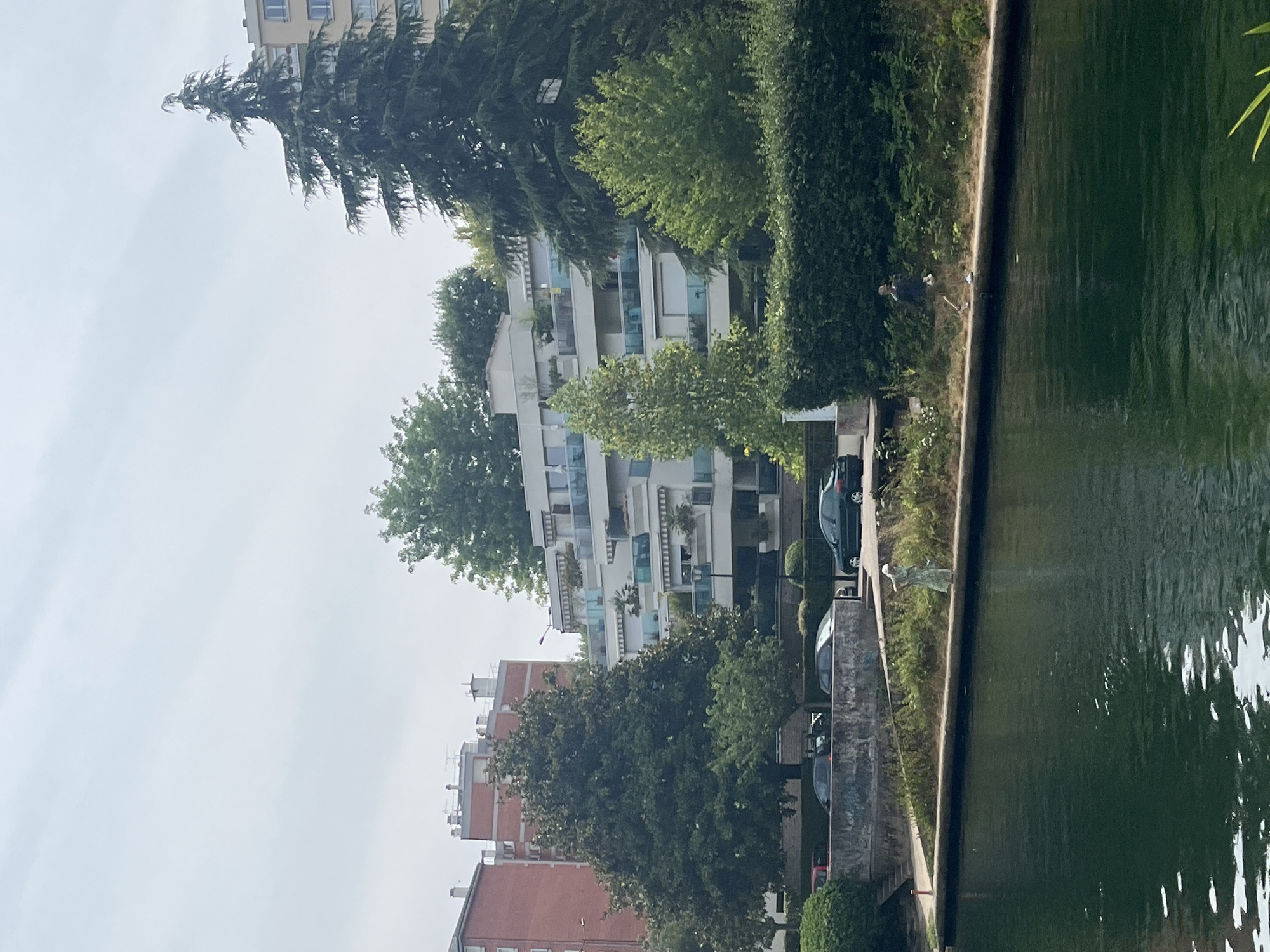 The image shows a canal in Joinville-le-Pont, France, with a paved walkway along its bank. On the walkway, a person wearing a light-colored dress is standing near some vegetation. Further down the path, another person wearing dark clothing is walking away from the camera. Behind the walkway, there are several cars parked along a street. Beyond the street, modern apartment buildings with balconies are visible, surrounded by trees. A large evergreen tree stands prominently on the right side of the frame, with its branches reaching towards the sky. The water in the canal is dark green and reflects the light from the sky.