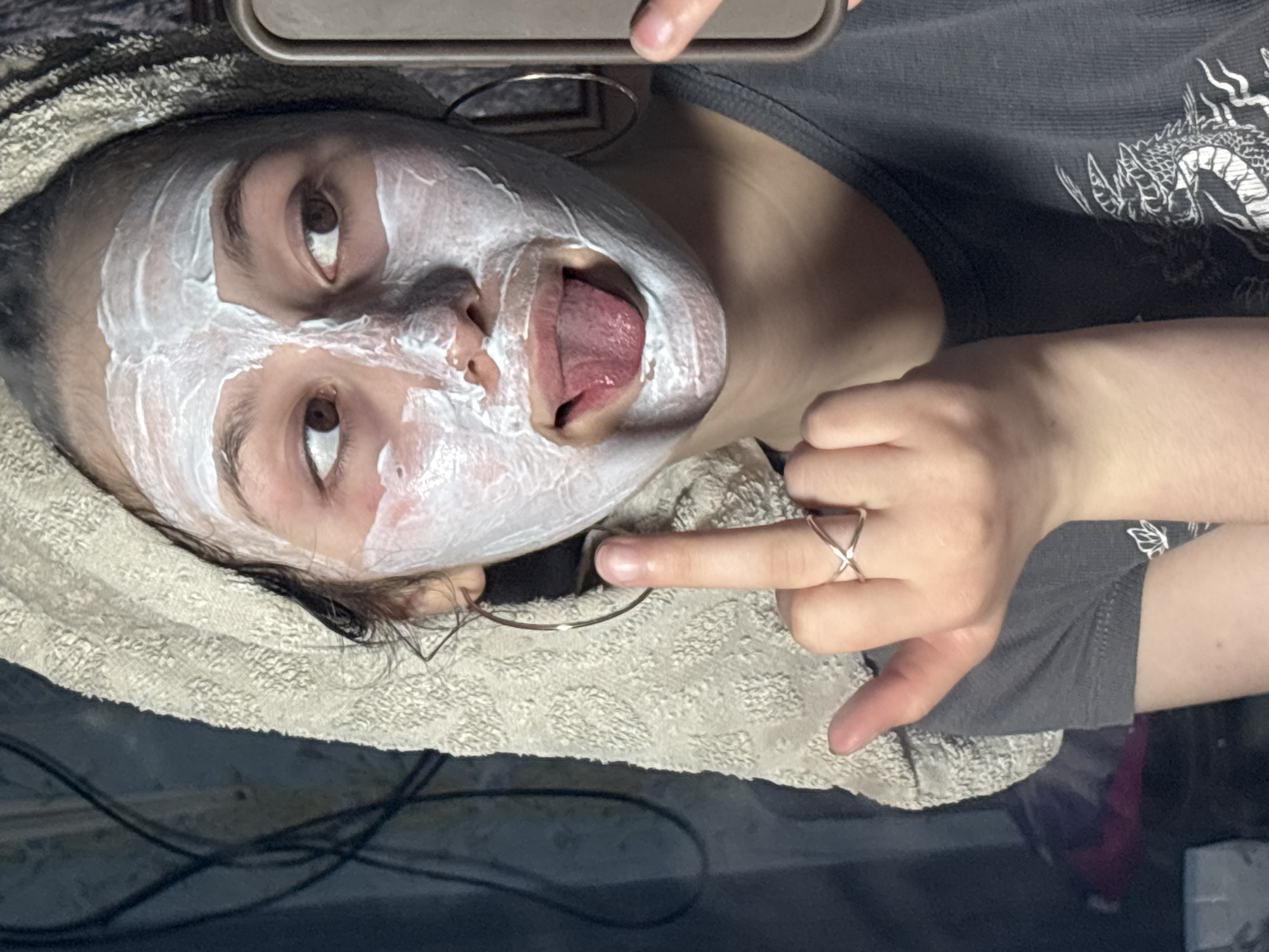 A young person with a white face mask and tongue extended is positioned on a light-colored towel. They are wearing a gray t-shirt with a dragon graphic. Their hand, adorned with a cross ring, is visible. An earring is also present. The environment includes dark-colored cords and furniture. The location, Фоки, Russia, is not directly discernible from the image.