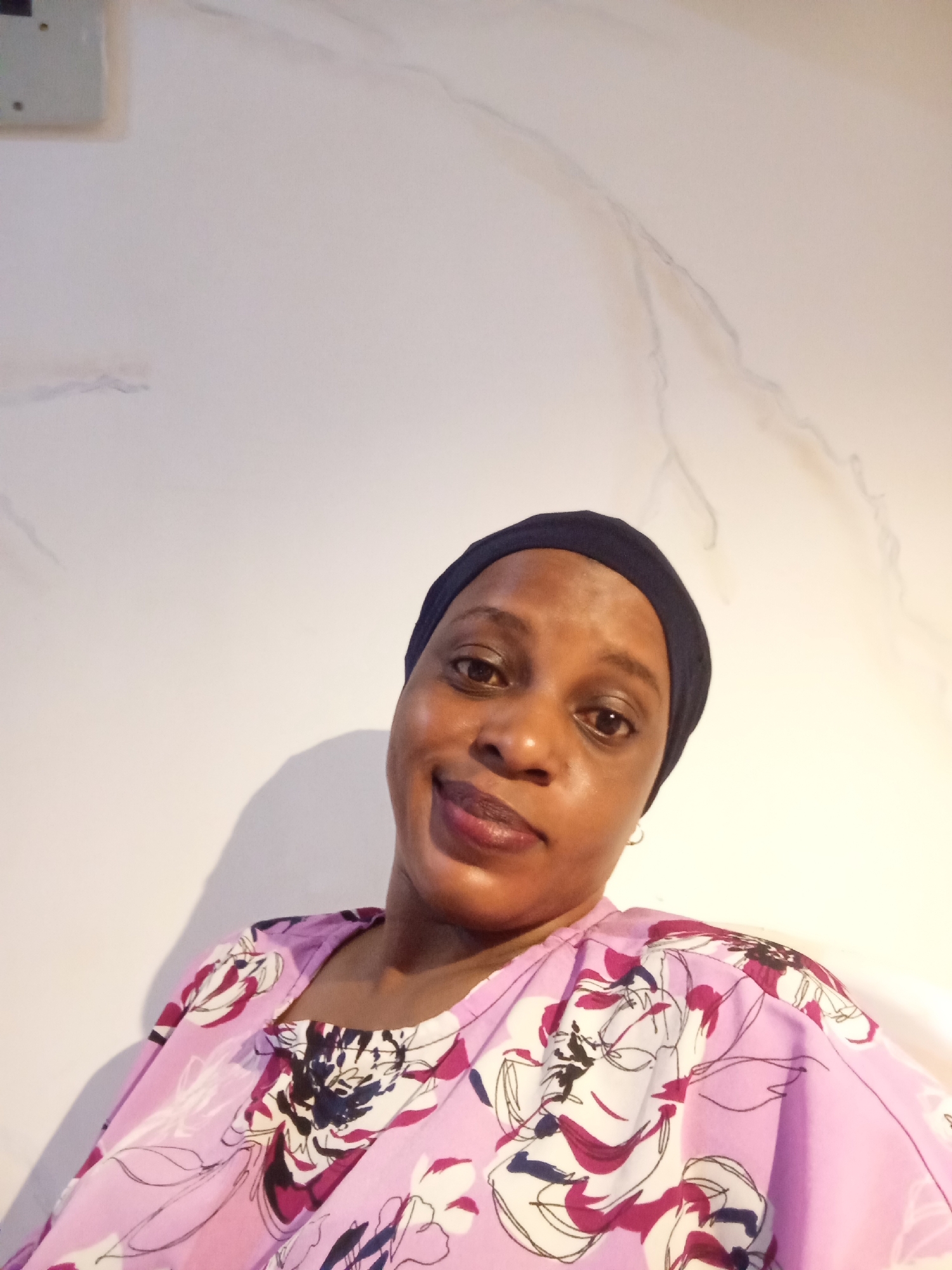 A medium close-up shot shows a person wearing a dark head covering and a pink floral blouse. The person is positioned against a light-colored wall, likely indoors. The photo seems to be taken from a slightly low angle. No specific details place the subject in Unguwar Rimi, Nigeria. There are no specific actions or interactions evident in the photo.