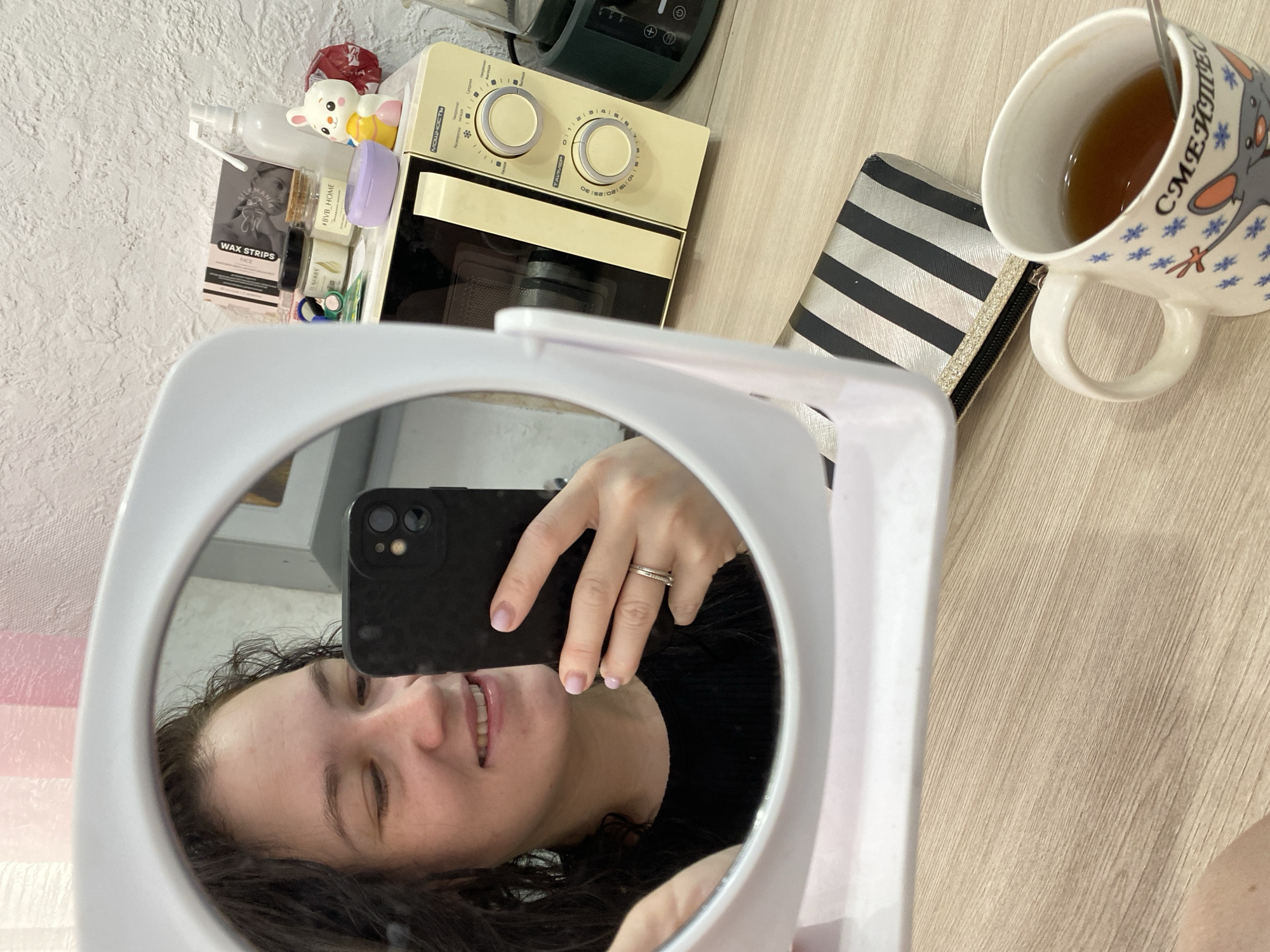 A person is taking a selfie in a mirror. The reflection shows the individual holding a black smartphone, their face close to the mirror, and their hand with a ring. The surrounding scene includes a mug with tea and a spoon, a striped pouch, a vintage-looking microwave oven, and various cosmetic or personal care items next to it. The background appears to be a wall with textured wallpaper and a wooden surface underneath. Based on the items present and the location inferred from web search results related to similar images, the location can be presumed to be in Chaikovsky, Russia, likely within a domestic setting.