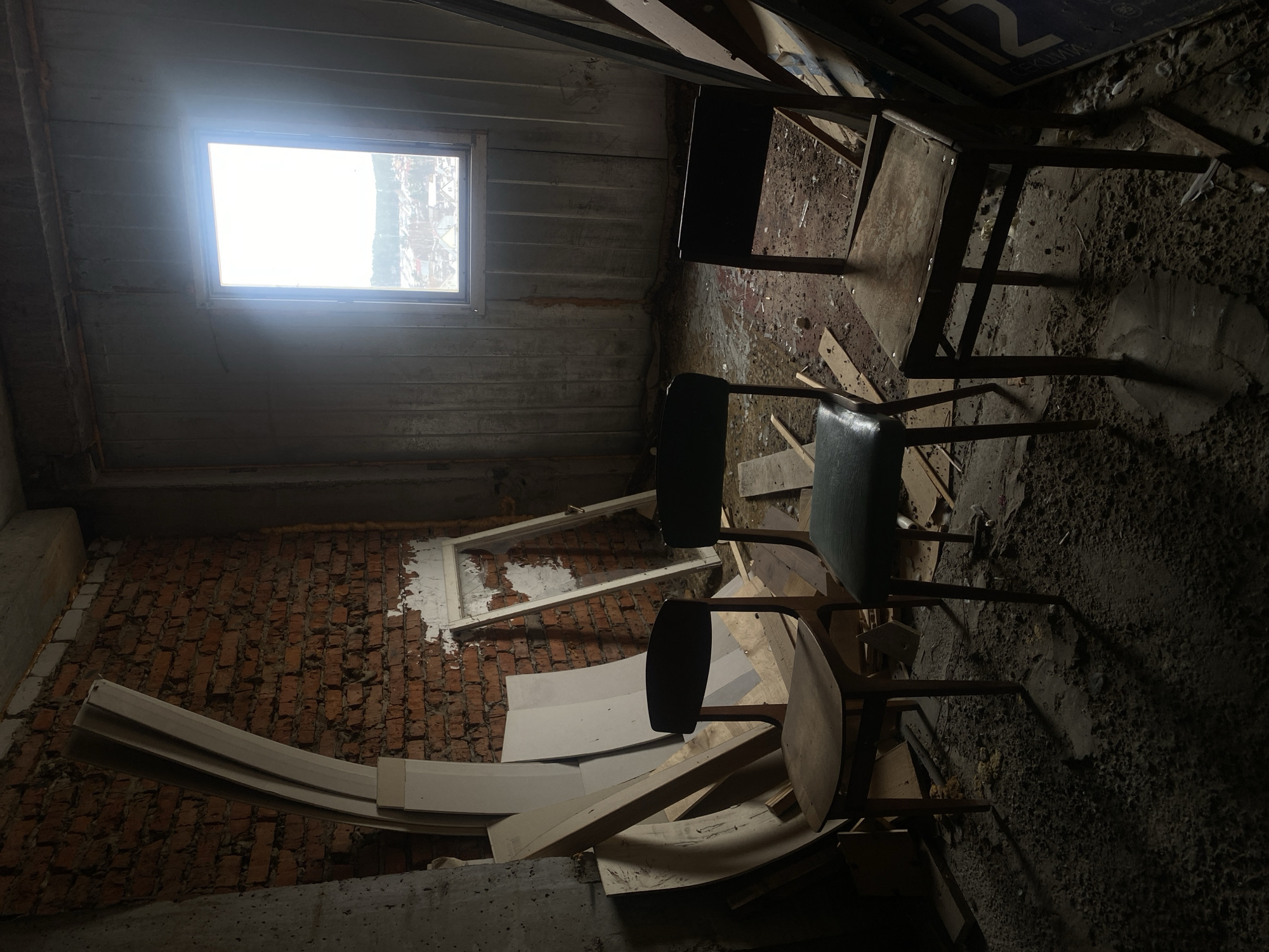 The image depicts the interior of a derelict space in Chaikovsky, Russia. A small window provides the primary light source. The walls show signs of decay, with brickwork partially exposed. Several chairs are present, positioned amidst debris and dirt. Discarded building materials, including pieces of trim and wood, are scattered on the floor. The scene overall conveys a sense of abandonment and disrepair.