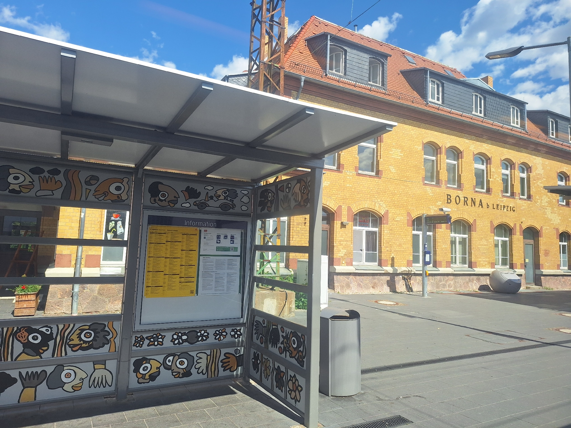 The image depicts a street scene in Borna, Germany, specifically in front of the train station. The foreground features a bus stop shelter with a gray metal frame and a white roof. The shelter's side panels are decorated with colorful, stylized illustrations of faces and abstract shapes. A yellow timetable is displayed within the shelter. To the right of the shelter is a gray trash can.
The background is dominated by a yellow brick building, which is the Borna train station. The building has a red-tiled roof with dormer windows. Above the entrance, the text "BORNA b LEIPZIG" is visible. A streetlamp is positioned to the right of the building. The ground is paved with gray tiles, and there are some shadows cast by the shelter and the building. The sky is blue with scattered white clouds.