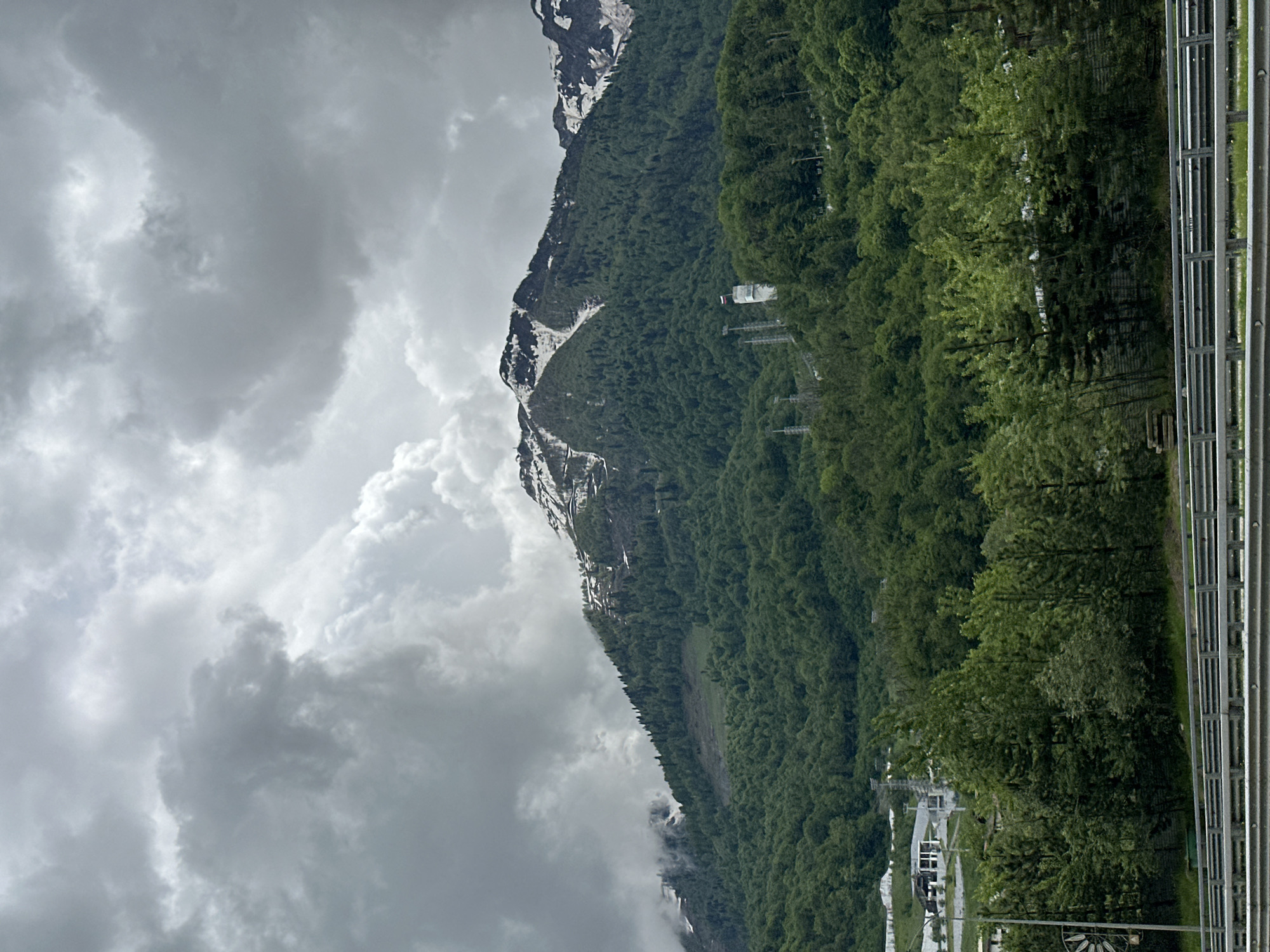 The image depicts a mountainous landscape likely taken in Krasnaya Polyana, Russia, judging by the terrain. The mountain slope is covered in dense green forest, interspersed with visible power line structures and occasional patches of snow at higher elevations. Clouds shroud the mountain peaks, obscuring the top of the mountain. The landscape is viewed from an elevated position, evidenced by the presence of a metal railing structure in the right of the image. At the foot of the mountain, some buildings are visible in a small settlement. The weather appears overcast and possibly stormy, given the heavy cloud cover.
