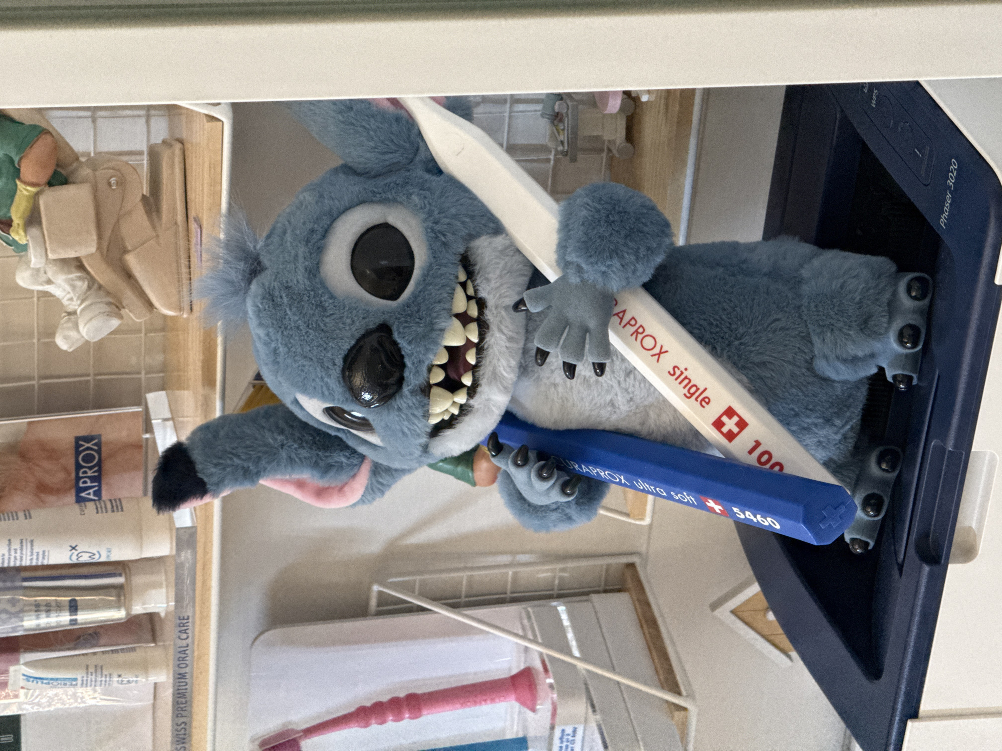 A stuffed blue alien character, resembling Stitch from Disney's Lilo & Stitch, is positioned in the foreground, clutching two large toothbrushes, one blue and one white. The blue toothbrush has text that reads "CURAPROX ultra soft 5460", and the white one reads "CURAPROX single 100". The character stands on a dark blue object, possibly a printer, with the words "Phaser 3020" visible on its surface. In the background, shelves are visible, displaying various oral care products, including tubes of toothpaste and toothbrushes. The overall scene appears to be an indoor setting, possibly a bathroom or a retail display area focused on dental hygiene. There is no indication of the location being in Мирновское сельское поселение, Ukraine.
