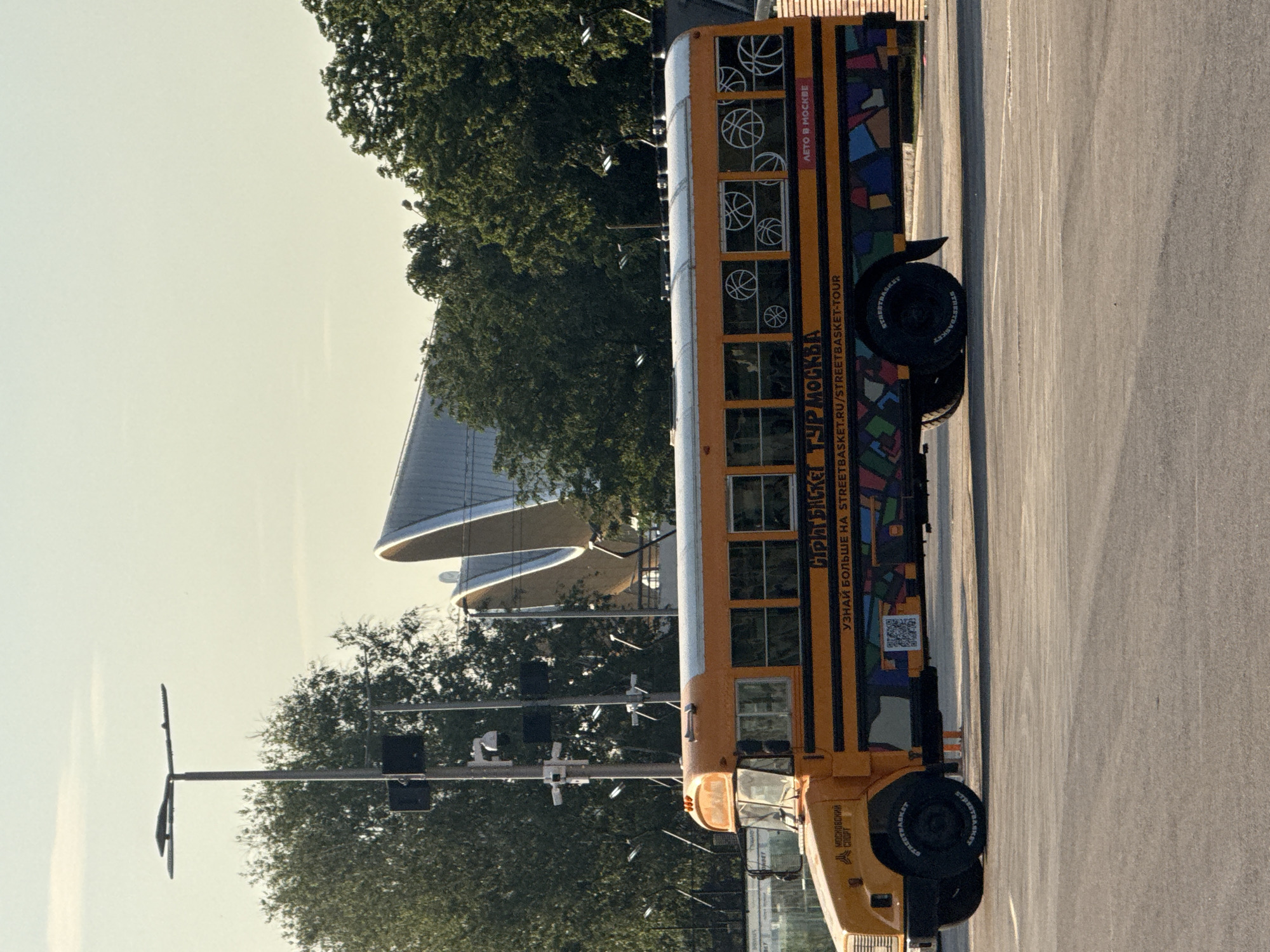 The image depicts an orange and yellow school bus parked in an outdoor paved area, likely a parking lot or street, in Moscow, Russia. The bus has black and yellow stripes along its side and is decorated with colorful geometric patterns and images of basketballs. Text on the bus reads "Streetbasket Tour Moscow" and provides a website address. In the background, a modern architectural structure with curved, metallic elements is visible, surrounded by lush green trees. A lamppost with multiple cameras is positioned to the left of the bus. The sky is a clear, pale yellow, suggesting it might be early morning or late afternoon. There are no people or animals visible in the image.