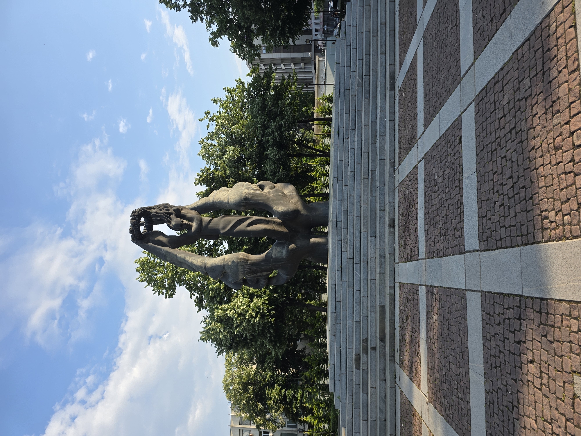 The image captures a public urban landscape, presented rotated 90 degrees clockwise from its natural orientation. In the center, a large, abstract bronze sculpture dominates the foreground, depicting two elongated, intertwined figures reaching upwards to hold a circular object, possibly a wreath or crown.
To the right of the sculpture, a wide, grand set of grey stone steps ascends, with individual steps clearly delineated. Further to the right, a paved area extends into the distance, made of reddish-brown cobblestones separated by smooth, lighter grey stone strips forming a grid pattern. Long shadows are cast across this paved surface, suggesting direct sunlight.
In the background, behind the sculpture and steps, a dense arrangement of lush green trees with varied foliage is visible, indicating a park or green space. Beyond the trees, several multi-story urban buildings can be seen under a bright blue sky dotted with scattered white clouds.
The clear sky and strong shadows suggest it is daytime, likely morning or late afternoon. No visible text is present in the image.
