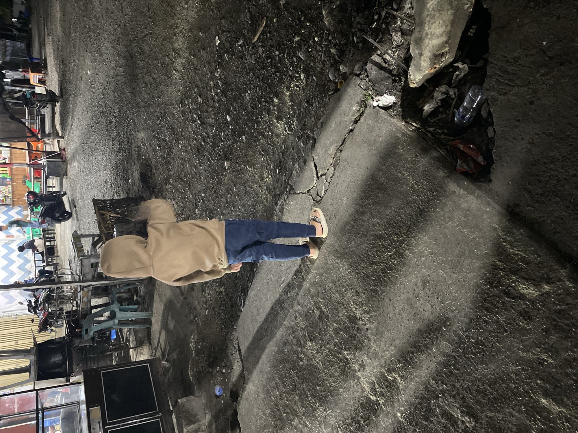The image depicts an outdoor scene at night, likely in a public area within Banda Aceh, Indonesia. A young child, viewed from behind, stands on a cracked concrete surface. The child is wearing a tan hooded sweatshirt and blue pants. Their right arm is raised, possibly gesturing.
The surrounding environment consists of gravel and broken concrete. In the background, several motorcycles are parked. To the left, a dark metal counter or display case is visible. Further back, there are structures that appear to be food stalls or market stands, with tables and chairs, and other individuals are present, including one person wearing a hijab. The ground is littered with debris, including a plastic bottle and what appears to be crumpled paper or fabric. The lighting suggests artificial light sources, typical of a nighttime urban setting. The overall impression is that of a casual gathering or market area after dark.