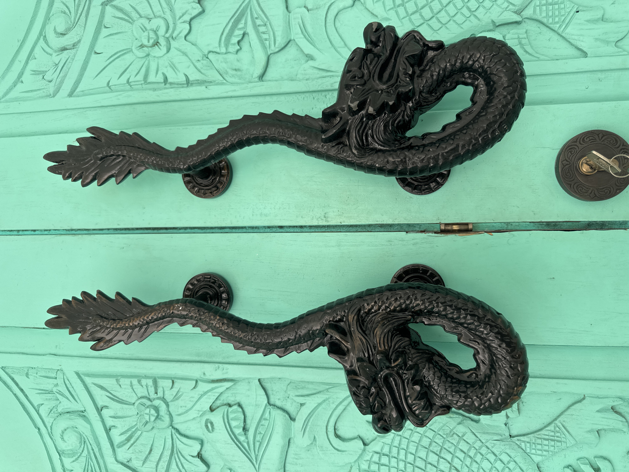 This close-up shot features two identical, ornate dark-colored dragon-shaped door handles set against a vibrant teal-painted wooden door, showcasing an architectural detail from Kuta Selatan, Indonesia.
The main subjects are the two prominent handles, crafted from a dark, possibly cast metal with a matte finish, depicting stylized dragons. Each dragon handle is mounted horizontally. The dragon's head, intricately detailed with scales, eyes, and a textured mane or frill, forms one end, serving as an elaborate grip. The body extends from the head, forming a curved handle with a scaly texture, and terminates in a pointed, leaf-like tail structure. Each handle is secured to the wooden door by two circular, dark metal bases with decorative fluting.
The backdrop is a wooden door, painted a striking, bright teal or turquoise color. The door itself is intricately carved with traditional relief patterns, possibly floral or abstract designs, which are partially filled with the paint, indicating texture and depth. The paint shows slight wear in places, revealing some of the wood grain underneath.
To the right of the upper handle, a circular, dark metal door lock mechanism is visible, featuring a key inserted into its keyhole. The lock's escutcheon is adorned with subtle, embossed patterns. A thin, horizontal metal strip, likely a hinge component or part of the door's construction, is visible between the two main wooden panels at the bottom center of the frame. The lighting suggests a bright environment, casting gentle reflections on the handles. There is no visible text in the image.