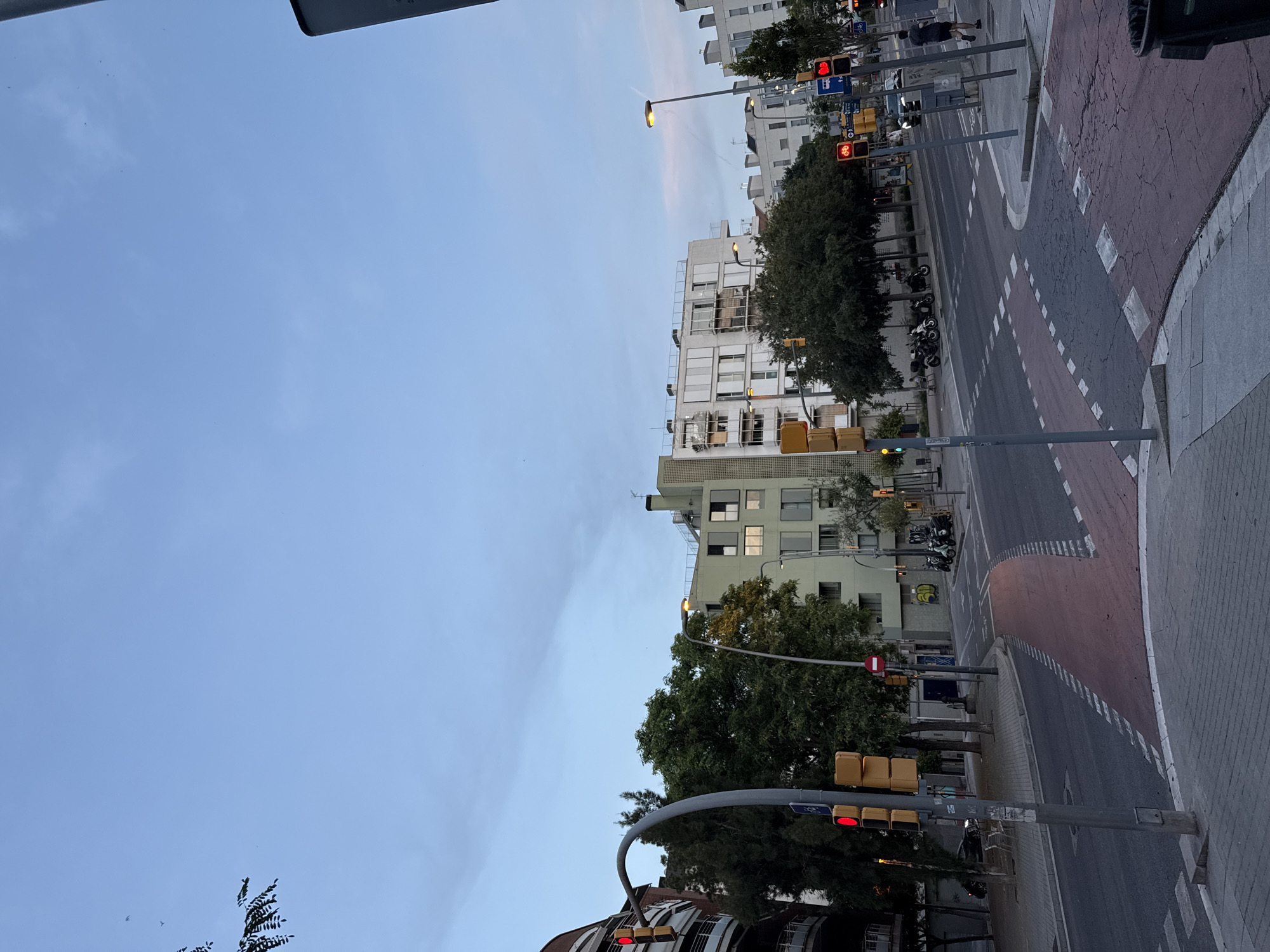 The image depicts a street scene in Barcelona, Spain, likely at dusk or dawn due to the soft, diffused light. The sky is a pale blue with some wispy clouds. The perspective is from an elevated position, looking down a street that appears to be a major intersection.
Dominating the scene are several multi-story buildings, typical of urban European architecture, with light-colored facades and numerous windows. Trees line the street, their dark green foliage contrasting with the buildings. The street itself is paved and marked with pedestrian crossings and traffic lanes. Several traffic lights are visible, with some showing red signals, indicating a pause in traffic. A few motorcycles are parked along the sidewalk.
There are a few people visible on the street, some walking and others possibly waiting at the traffic lights. One individual in dark clothing is seen walking away from the camera.
Notable details include the street markings, which suggest a dedicated bike lane in some sections. The presence of traffic lights and the general urban layout strongly indicate a city environment. The lighting suggests it is either early morning or late evening. No specific text is clearly legible in the image, though some signs are present on poles. The overall mood is calm and quiet, typical of a city at the transition between day and night.