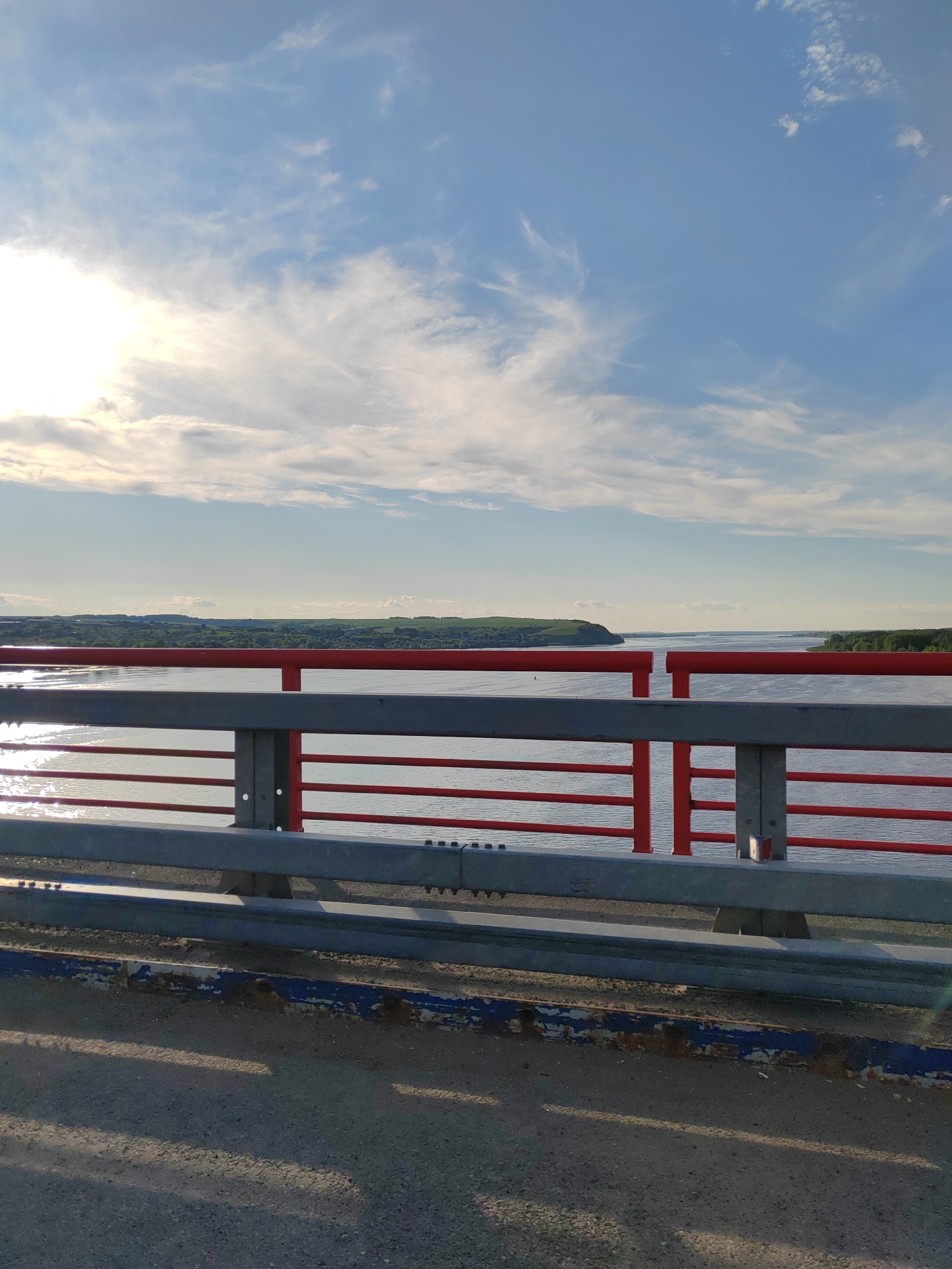 The image presents a daytime view from a bridge overlooking a vast body of water, likely a river or large lake, framed by distant hills and land on the horizon. In the foreground and mid-ground, the bridge's safety railing is prominent, consisting of bright red horizontal bars supported by grey/silver vertical posts and lower beams. Below the railing, at the very bottom of the frame, is a worn blue and white barrier along the edge of the asphalt road surface, showing signs of rust and peeling paint.
The water below the bridge reflects the strong sunlight from the left, creating bright, shimmering streaks across its surface. The distant landscape on either side of the water features green, undulating hills and what appear to be forests or cultivated fields stretching towards the horizon. The sky above is a clear blue, dotted with scattered white, wispy clouds. The sun, partially visible on the upper left, casts a bright glow and some lens flare, indicating a sunny day, likely morning or late afternoon given the angle of the light and the long shadows on the bridge's surface. No people or specific activities are visible, presenting a serene, expansive landscape captured from a moving or stationary vehicle on the bridge. No visible text is present in the image.