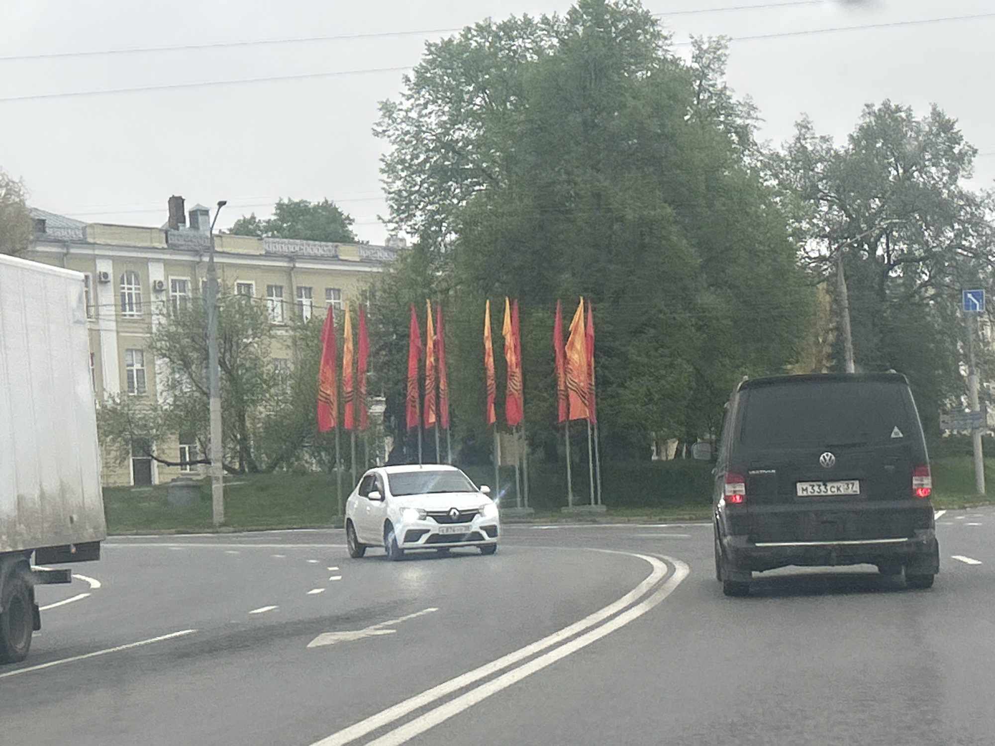 The image depicts a street scene in an urban environment. A white Renault Logan is approaching a roundabout with several tall red and orange flags on its perimeter. Behind it, a black Volkswagen van with license plate M333CK77 is also navigating the roundabout. To the left, a white truck is partially visible. In the background, there is a light yellow, multi-story building and a large tree. The road is wet, suggesting recent or ongoing rain. A blue road sign is visible on the right side of the road.
