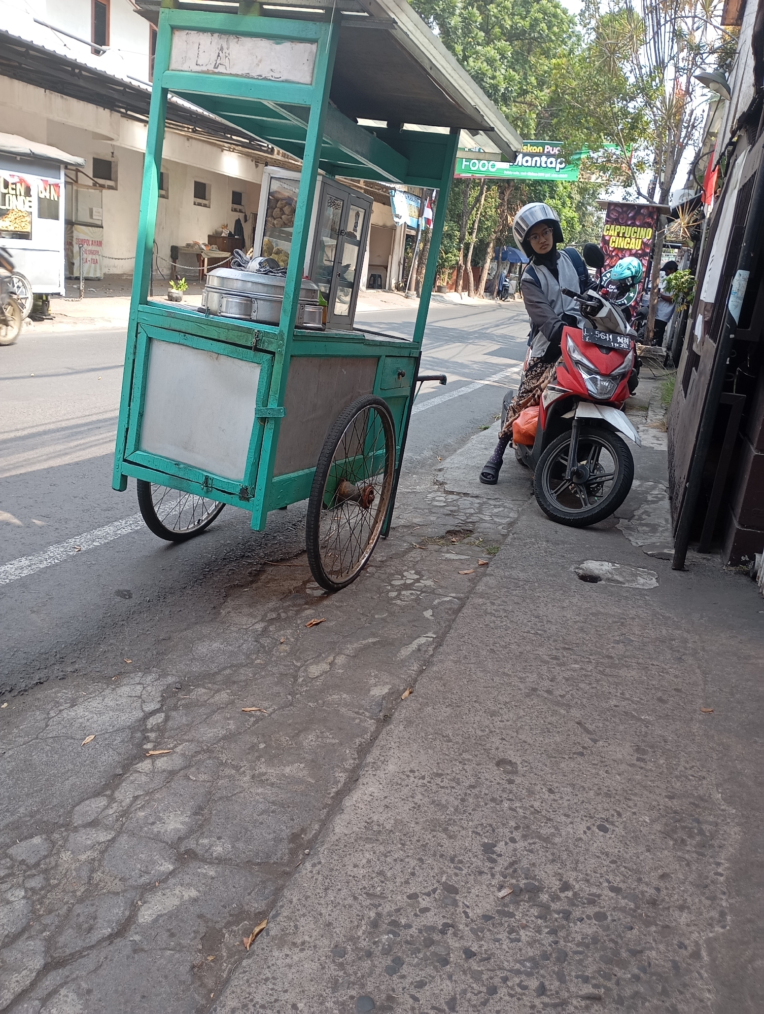 A young Indonesian woman wearing a gray jacket, black hijab, helmet, and glasses is seated on a red and white motorcycle in the city of Malang, Indonesia. She is on a street, next to a green and white food cart with wheels. The food cart is filled with stainless steel containers and has glass displays. A green sign with white text that reads "FOOD Mantap" is visible above the cart. The street is paved with asphalt and concrete, showing signs of wear and tear. Trees and buildings are visible in the background, indicating an urban environment. The lighting suggests it is daytime, with sunlight casting shadows on the ground.