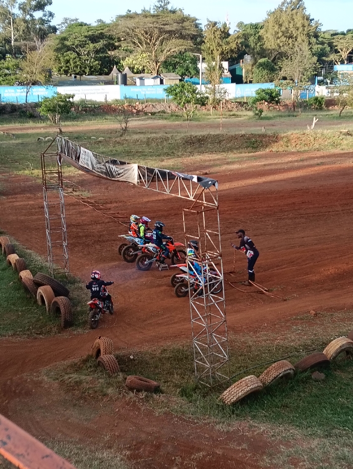 A dirt track with several young motocross riders lined up for a race start. A race official holds a starting gate. Tires line the edges of the track. One lone rider pulls onto the track from a small hill, away from the other riders. A metal starting gate structure towers over the scene. In the background is a mix of greenery, a water tank, and a blue wall. The sky is bright and clear.
