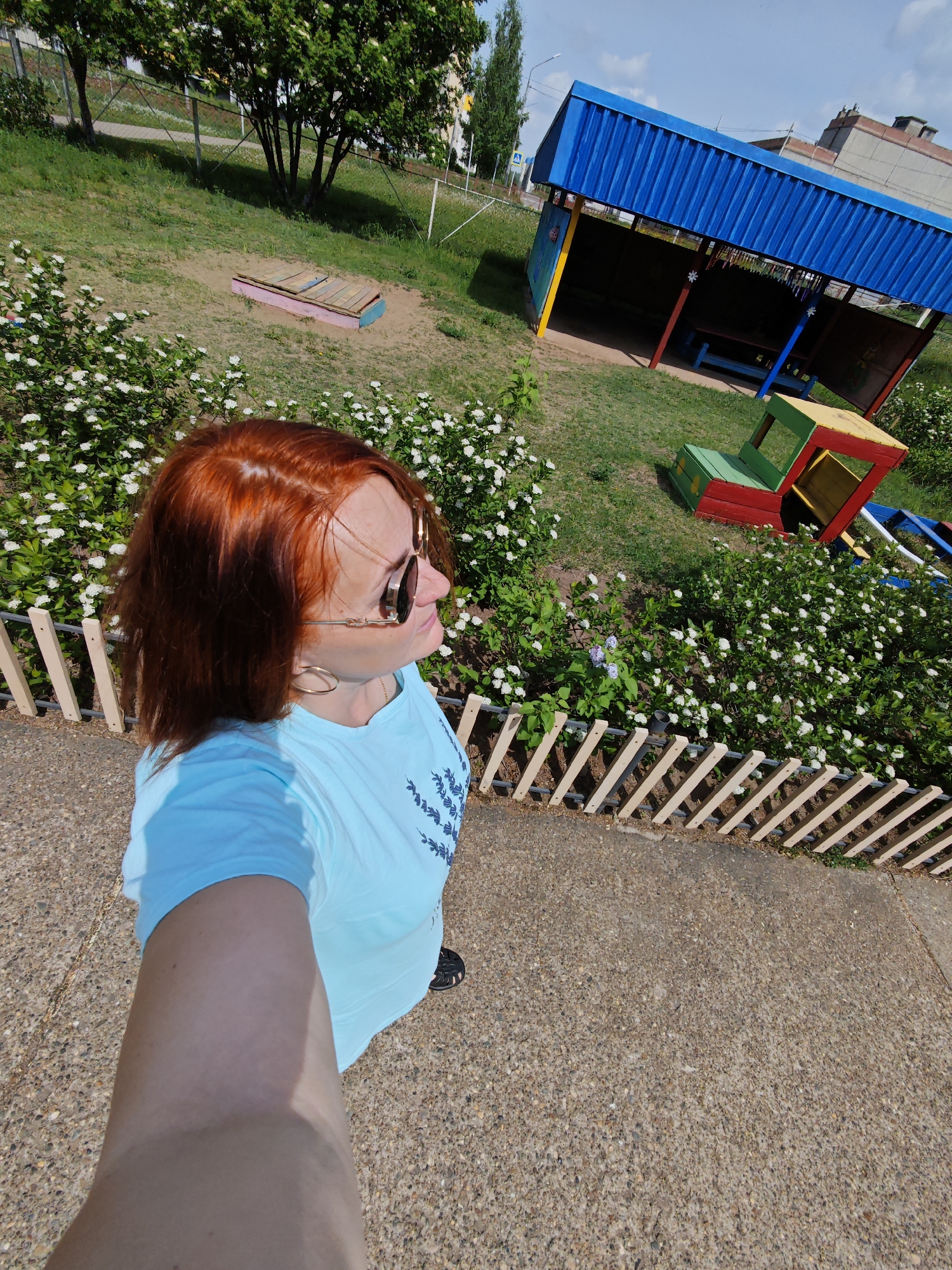 A woman with red hair and sunglasses, wearing a blue t-shirt and gold hoop earrings, takes a selfie outdoors on a sunny day. She stands on a concrete surface next to a low wooden fence, with flowering bushes behind it. In the background, there's a sandbox, a blue and yellow structure, a colorful play vehicle, and greenery, suggesting a playground or park. The location is identified as Марковский, Russia.