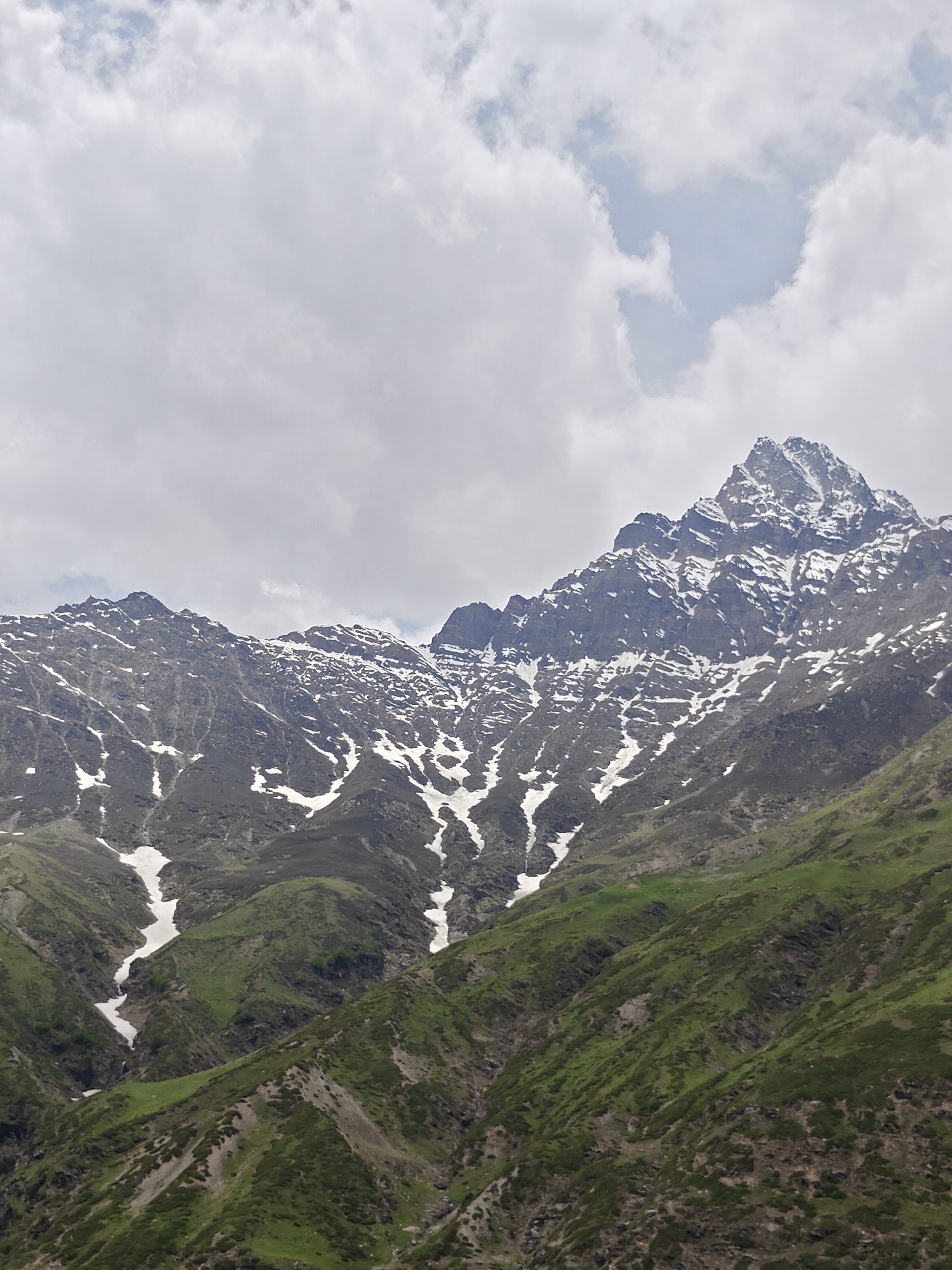 The image displays a majestic mountain range under a cloudy sky. The peaks are rugged and covered with patches of snow, contrasting with the lush green slopes below. Valleys are visible, carved into the mountainside, with some areas still holding onto snow. The sky is overcast with large, diffused clouds, suggesting it might be a cooler day or that rain could be imminent. The scene is devoid of any human presence or man-made structures, presenting a natural and wild landscape. The location is a remote mountainous area in India.
