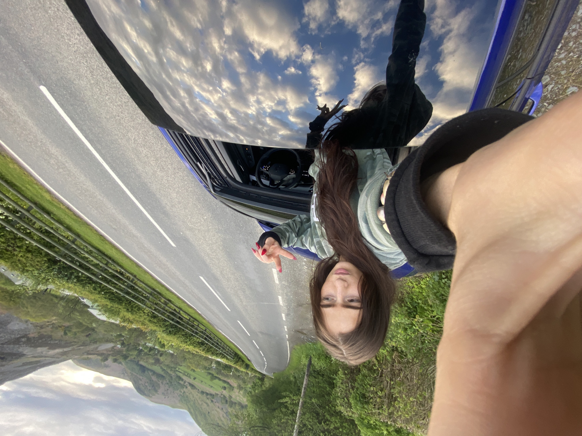 A selfie-style photo depicts a woman with long, dark hair, positioned next to a blue car on a road. She is making a peace sign with her left hand. The car hood reflects the sky with white clouds. The road stretches into the distance, bordered by green fields and trees. Mountains are visible in the background. Based on the visual cues of the landscape, this photo was likely taken near Llangynog, United Kingdom.