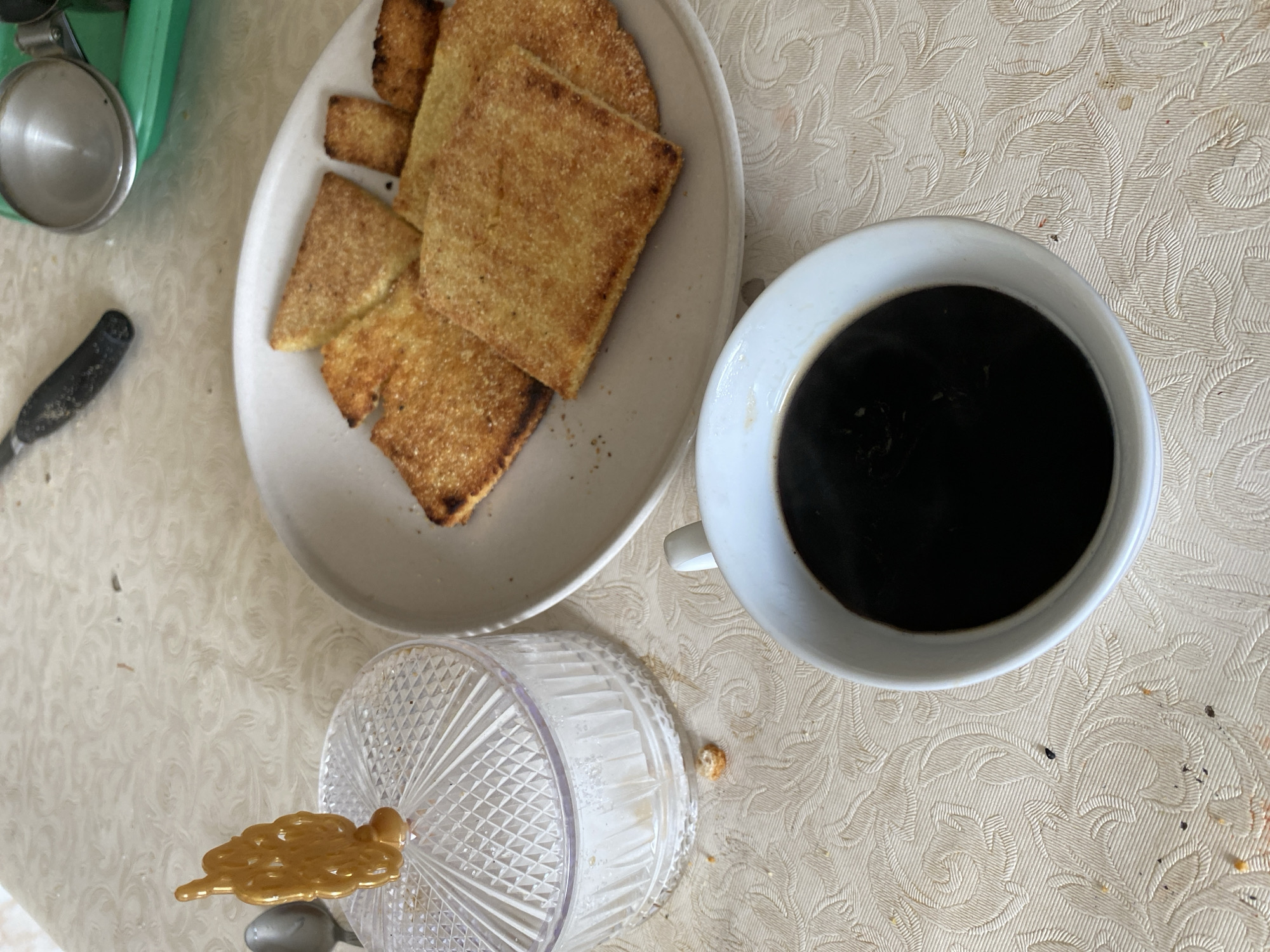 This is a high-angle, full shot of a table setting in Baraki, Algeria, featuring a cup of coffee, a plate of golden-brown, square-shaped crackers or biscuits, and a clear glass jar with a sugar scoop. The table is covered with a light-colored, patterned tablecloth. The coffee is in a white mug, with steam rising, indicating it's hot. The crackers are arranged on a white plate, some overlapping. The sugar jar is topped with a decorative, golden scoop. The background includes a glimpse of a utensil with a black handle and a green tray. The overall scene suggests a breakfast or snack setting.