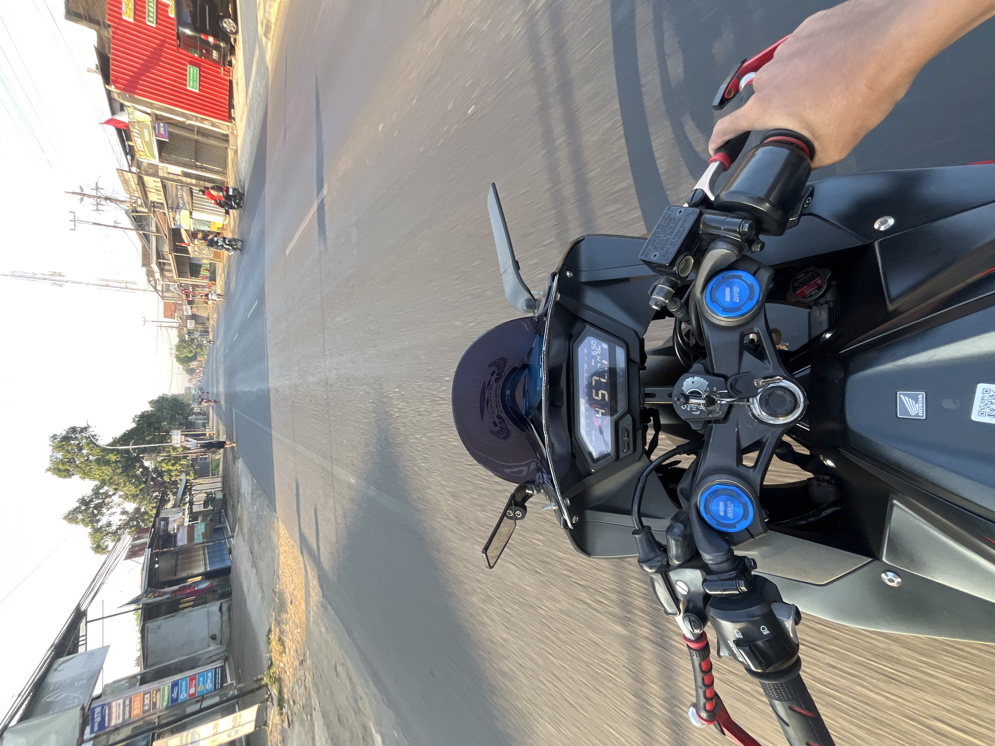 The image is a first-person perspective from a motorcycle traveling on a road in Banyuputih, Indonesia. The road is paved and has lane markings. On either side of the road are shops and buildings with various signs. Trees and utility poles are visible. Several other motorcycles are on the road, with some people riding them. The sky is clear and the sun is shining, casting shadows. The motorcycle's dashboard is visible, showing a speed of 45.7 km/h and a time of 6:50. The rider's hands are on the handlebars, with one hand operating the throttle. The motorcycle is black with red accents on the handlebars. The overall scene depicts a typical day on a road in a semi-urban area of Indonesia.
