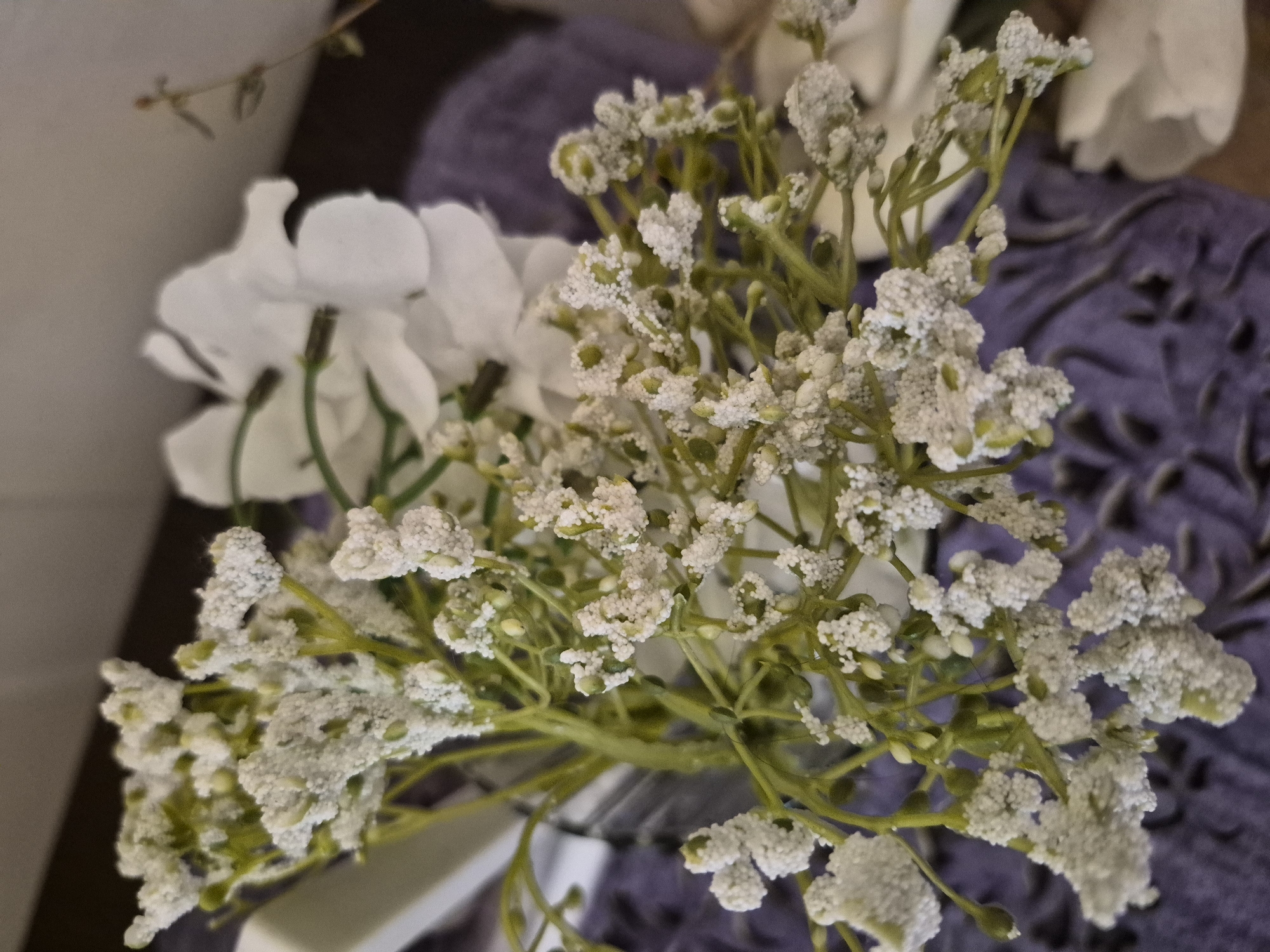 This is a close-up, eye-level shot of an artificial floral arrangement, likely displayed indoors. The primary subjects are two types of artificial flowers. In the foreground and sharp focus, there is a cluster of small, delicate white flowers resembling baby's breath, characterized by a distinct granular, possibly frosted or flocked texture on their petals. These flowers are attached to thin, bright green stems.
Behind and to the left of the textured white flowers, larger, smooth white artificial flowers are visible, appearing slightly out of focus. The background on the right features a soft purple fabric or surface with an embossed or patterned design, also out of focus. On the left side, a plain, light grey or beige surface, possibly a wall, is visible. In the upper left corner, a slender, dark brown branch with a few tiny green leaves extends into the frame.
The image has a shallow depth of field, drawing attention to the textured white flowers. There is no specific activity taking place, as it's a still life. The lighting appears soft and diffused, without harsh shadows. No text is visible in the image. While the location context is given as Yerevan, Armenia, there are no specific visual cues within the image to confirm this.