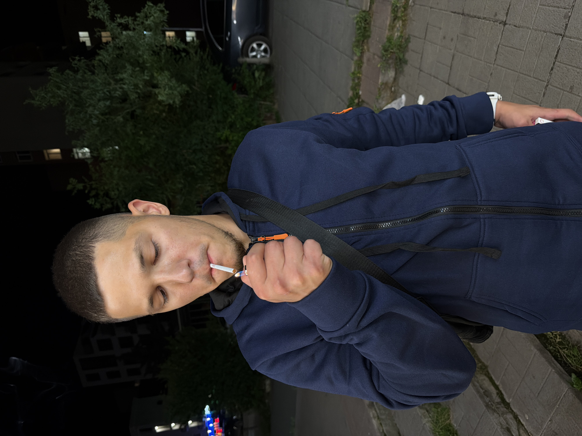 A young man is lighting a cigarette at night outdoors. He is wearing a dark blue hooded sweatshirt and a black strap across his chest. The background shows a dark urban environment with buildings and trees. The ground is paved. The man's face is illuminated by the lighter and the cigarette. The image does not contain enough information to determine the specific location in Sofia, Bulgaria.