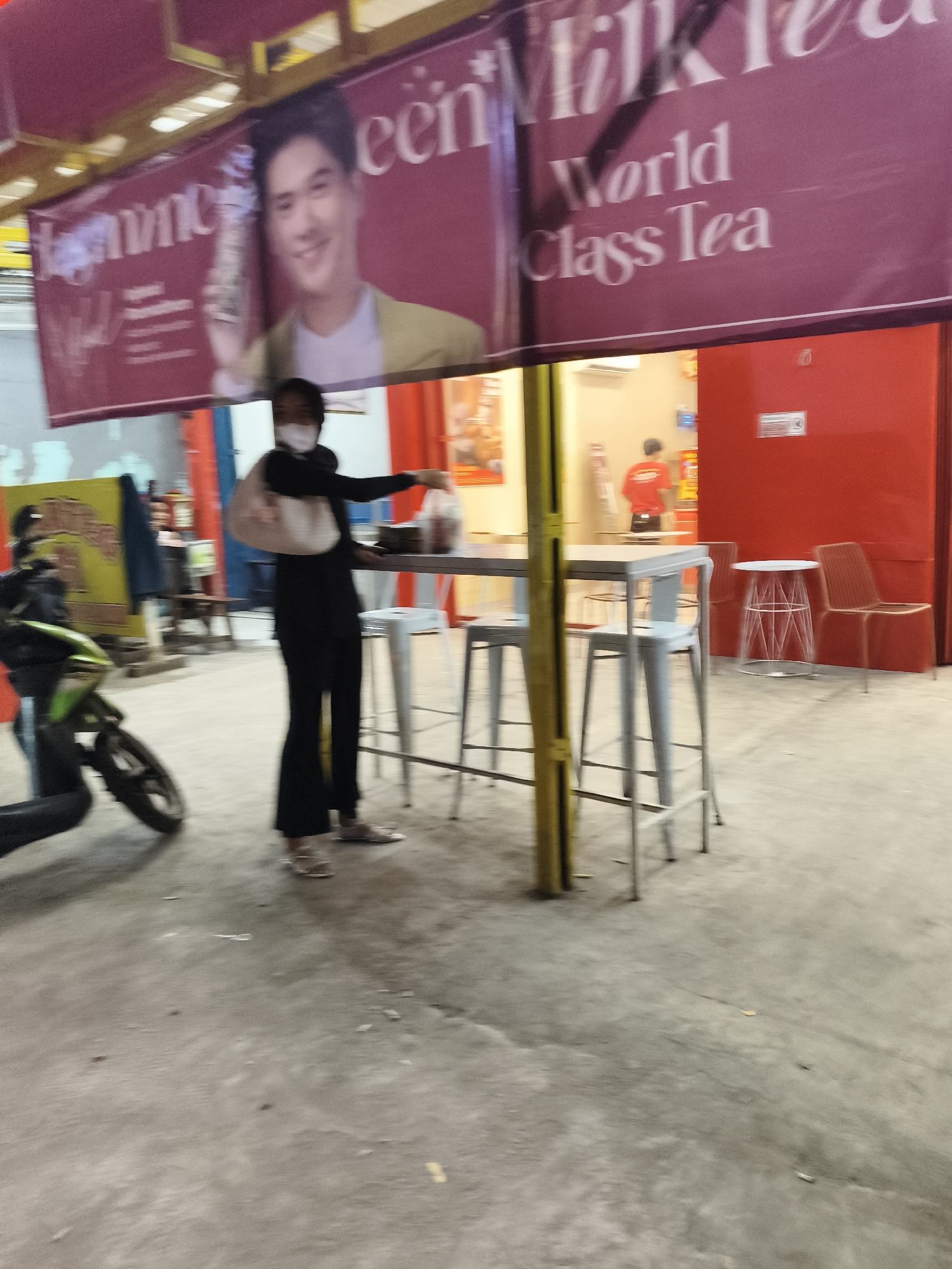 The image captures an outdoor setting, possibly a food stall or cafe, in South Tangerang, Indonesia. A prominent banner advertising "World Class Tea" with an image of a smiling man is displayed overhead. In the foreground, a person wearing a black outfit and a face mask is standing near a table with chairs, holding a plastic bag. A motorcycle is parked to the left, partially visible. The ground appears to be concrete, and the overall lighting suggests it might be evening or a dimly lit area. The background shows a red wall and some interior elements of a building, possibly indicating a shop or restaurant.