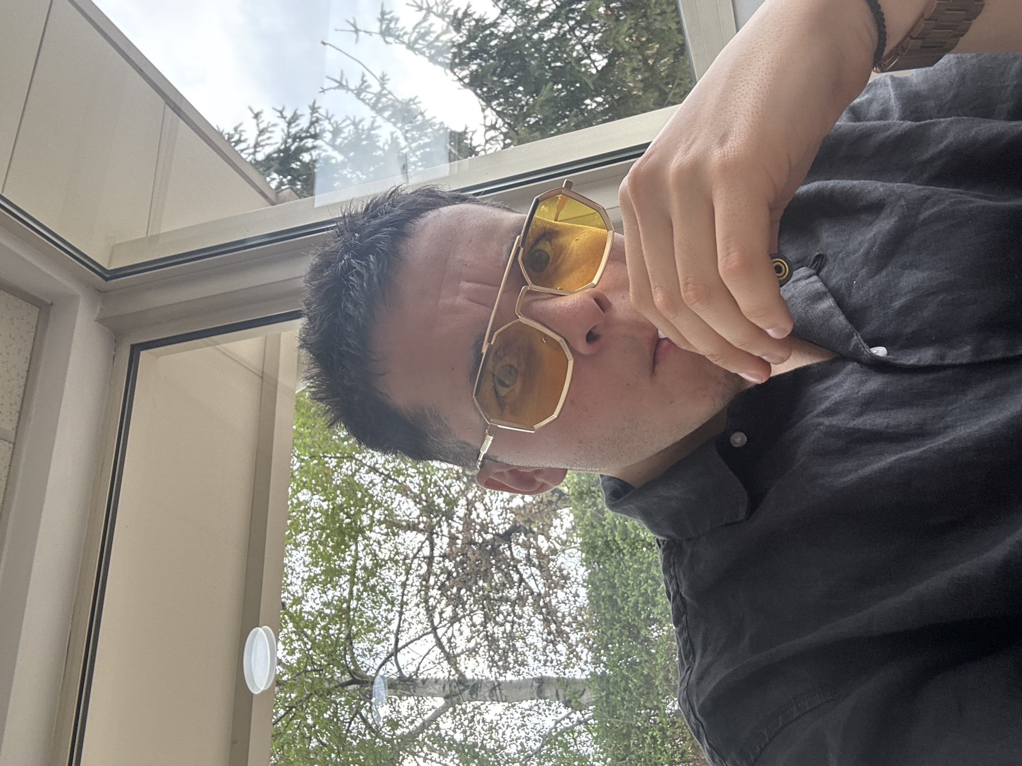 A young adult male is captured in a close-up, rotated portrait, appearing to be indoors and looking out through a large window or glass structure. He has short, dark hair and a neutral expression. He is wearing striking yellow-tinted, hexagonal or octagonal sunglasses with thin gold-colored metal frames; reflections of trees and an overcast sky are prominent on the lenses. A glimpse of his right eye is visible above the right sunglass lens.
He is dressed in a dark grey or black collared shirt, which appears to be made of a textured fabric like linen, with some buttons visible at the front. His right arm is raised, with his hand resting near his chest/shoulder area, partially covering a button on his shirt. On his right wrist, he wears a wooden watch with visible links and a slender black braided bracelet.
The background shows a bright, overcast sky and various trees with both bare branches and green foliage, seen through clear glass panels. The structure visible around the person includes light-colored or white window frames and architectural elements, suggesting a modern building, a conservatory, or an enclosed patio. A small white circular object, possibly a suction cup or decorative element, is attached to one of the vertical window frames. The bright natural light indicates it is daytime. The location is Sofia, Bulgaria. No text is visible in the image.