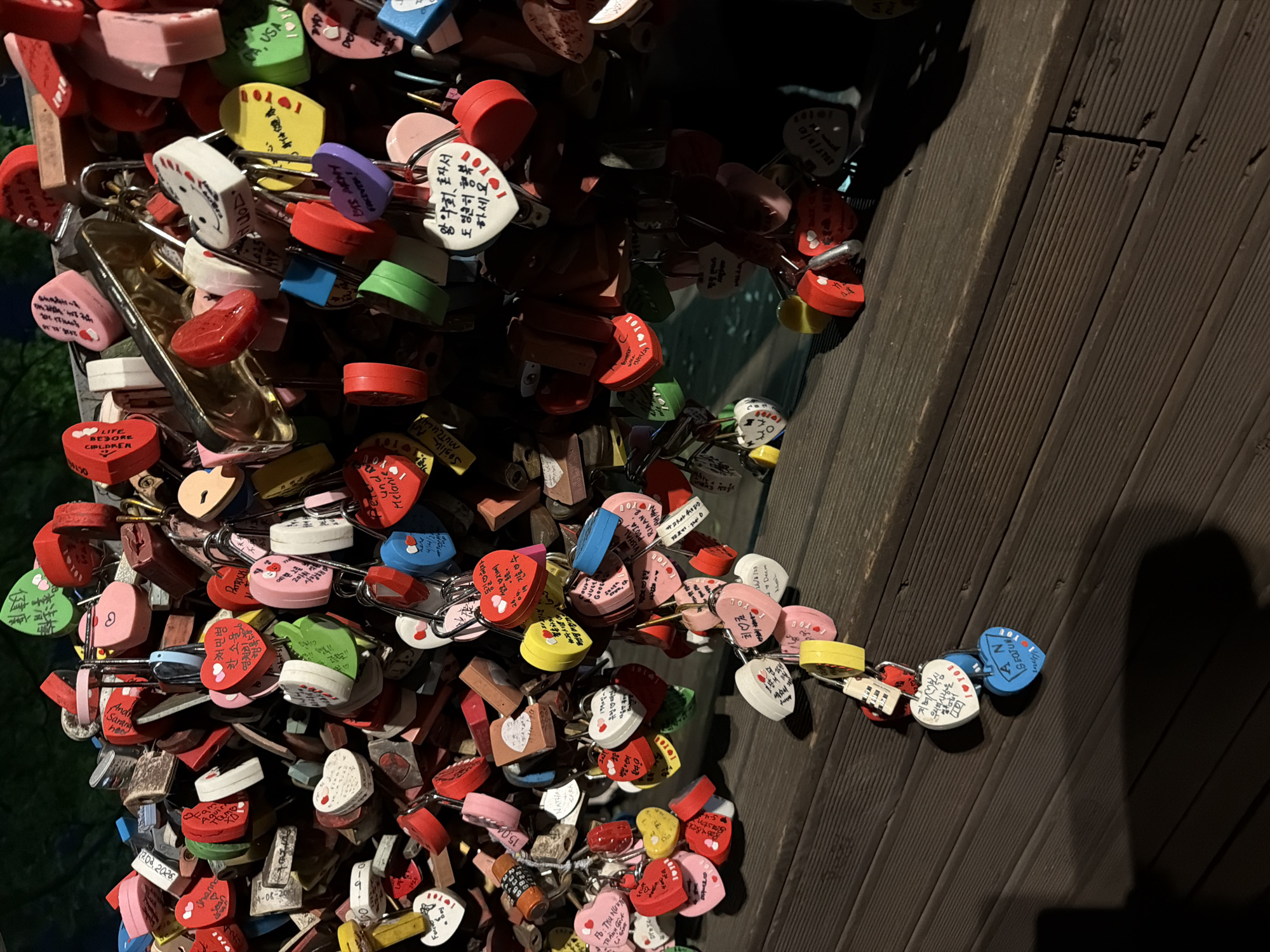 A mass of colorful love locks are attached to a metal railing or structure adjacent to a dark, wooden wall, likely a viewing platform. The locks are mostly heart-shaped or rectangular with rounded edges and are painted in red, white, pink, yellow, green, and blue, with some featuring messages or names written on them in various languages. The scene appears to be at night or in low-light conditions, with the locks illuminated by artificial light. The location is likely N Seoul Tower in Seoul, South Korea, as these love lock installations are commonly found there.