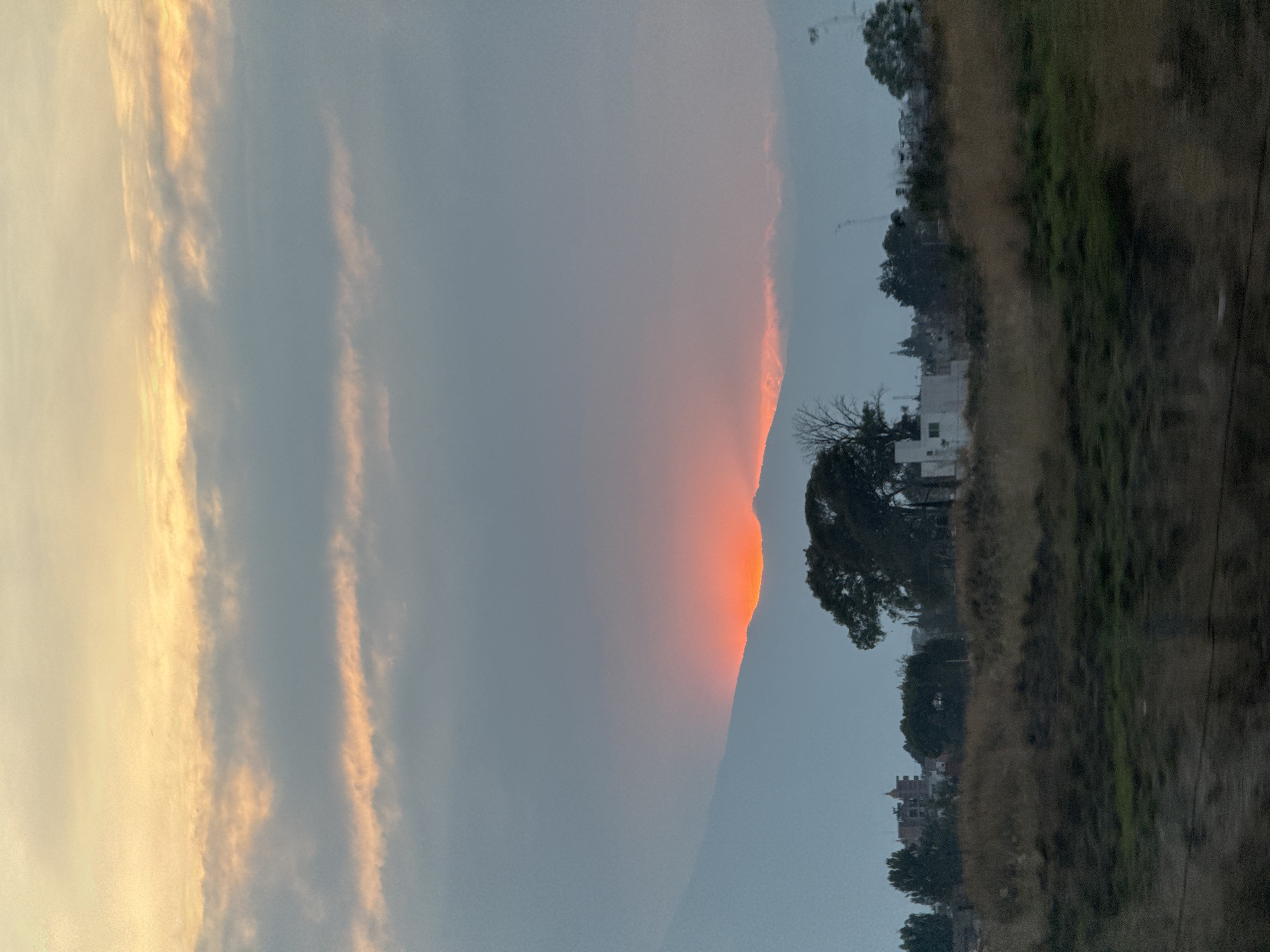 The image depicts a scene in San Bernardino Tlaxcalancingo, Mexico. A mountain is visible with a bright orange light striking its peak, contrasting with the dull sky. To the right, a landscape of vegetation, trees, and houses is visible. The sky is pale with striated clouds, suggesting either sunrise or sunset.