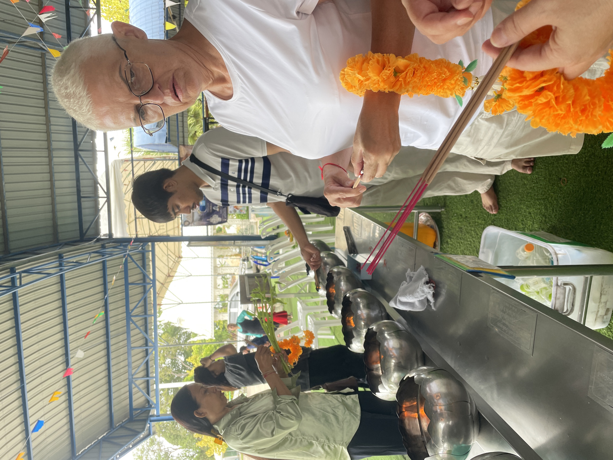 The image depicts a scene at a temple in Bang Sare, Thailand. In the foreground, a woman with short, blonde hair and glasses is holding a lit incense stick, preparing to place it in a large metal bowl. She is wearing a white t-shirt and light-colored pants, and an orange marigold garland is draped around her wrist. Beside her, a woman in a light green shirt is holding a bouquet of orange marigolds and green leaves, and another woman in a dark t-shirt is also holding a similar bouquet. In the middle ground, a young man in a striped shirt is standing by a table, also appearing to be involved in the ritual. In the background, there are rows of white plastic chairs and glimpses of trees and buildings, suggesting an outdoor, covered area typical of a temple complex. The metal bowls on the table appear to be filled with burning incense or oil. Decorative flags are strung across the ceiling structure.