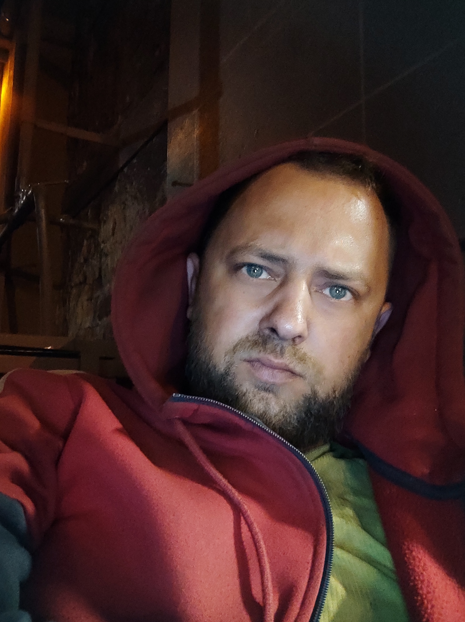 The image is a close-up, low-angle selfie of a man wearing a red hooded sweatshirt and a light green shirt. He has light skin, blue eyes, a beard, and is looking directly at the camera with a neutral expression. The background is dark and appears to be an outdoor urban setting, possibly a street or alleyway, with visible stone or brickwork and some structural elements that suggest a building. There is a yellow light source visible on the left side of the frame. The image was taken at night, as indicated by the darkness of the background and the artificial lighting. The specific location in Salzgitter, Germany, cannot be determined from the visual information present in the image.