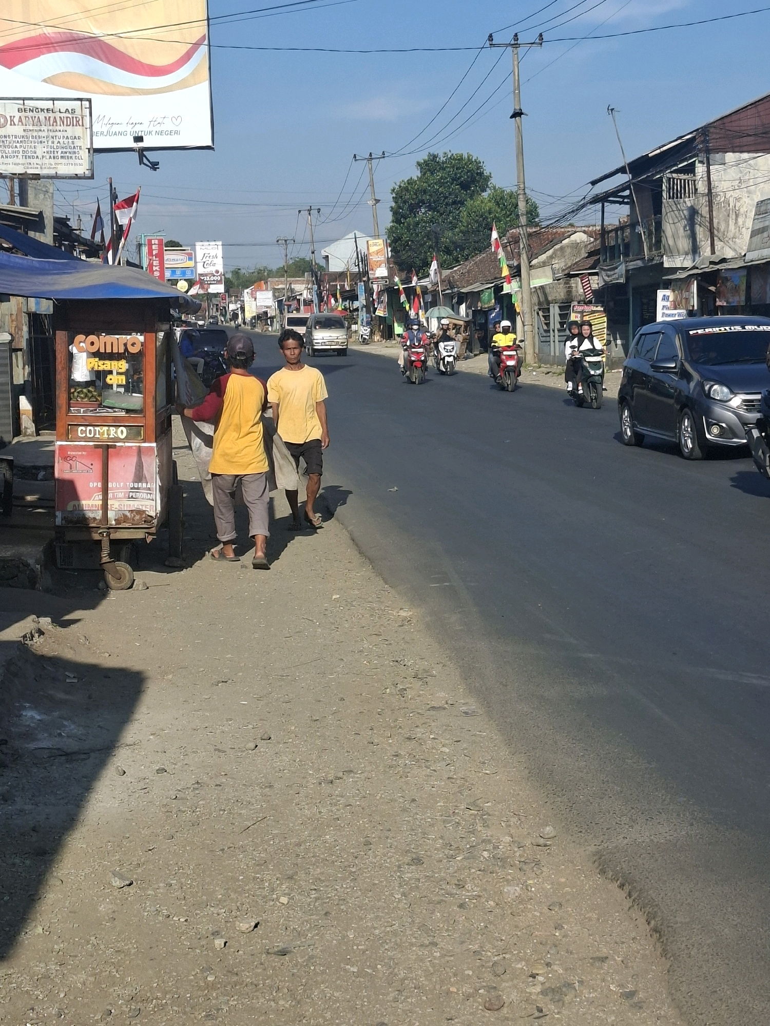 The image captures a street scene in Lembursawah, Indonesia, characterized by a clear blue sky and bright sunlight casting distinct shadows. The foreground shows a dirt and gravel path bordering an asphalt road. On the left side of the road, a small food cart with a "Comro Pisang" sign is visible, with two men standing near it. One man, wearing a yellow t-shirt and dark shorts, is walking away from the cart, while another man, in a red and yellow shirt and grey pants, is facing the cart, holding a large piece of fabric.
The road itself has a moderate amount of traffic, with several motorcycles and a dark-colored car moving. Flags, including the Indonesian flag, are displayed along the roadside, and various signs are mounted on buildings and poles. The buildings lining the street appear to be a mix of residential and commercial structures, some with corrugated metal roofs and weathered facades. Power lines and utility poles are prominent features of the urban landscape. The overall impression is that of a bustling street in a developing area, with everyday activities unfolding under the midday sun.