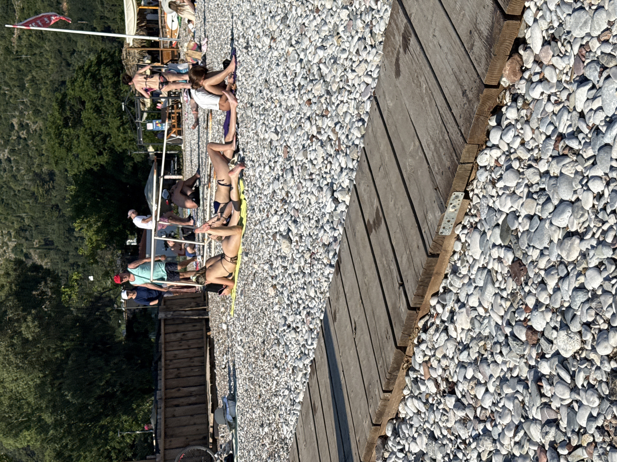 The image depicts a sun-drenched beach scene in Budva, Montenegro. A group of people are scattered across a pebble-covered shore, some sunbathing on towels while others appear to be relaxing. A wooden boardwalk runs diagonally across the frame, separating the pebble beach from a grassy, tree-covered hillside in the background. Several beach umbrellas and a small stand are visible further up the beach, suggesting a developed tourist area.
The time of day appears to be late afternoon, with long shadows cast by the wooden boardwalk. The weather is clear and sunny, typical for a summer day in Montenegro. The people are dressed in swimwear, indicating they are at the beach for leisure activities such as sunbathing or swimming. There is a red and white flag flying from a flagpole on the left side of the image. The overall mood seems relaxed and summery.