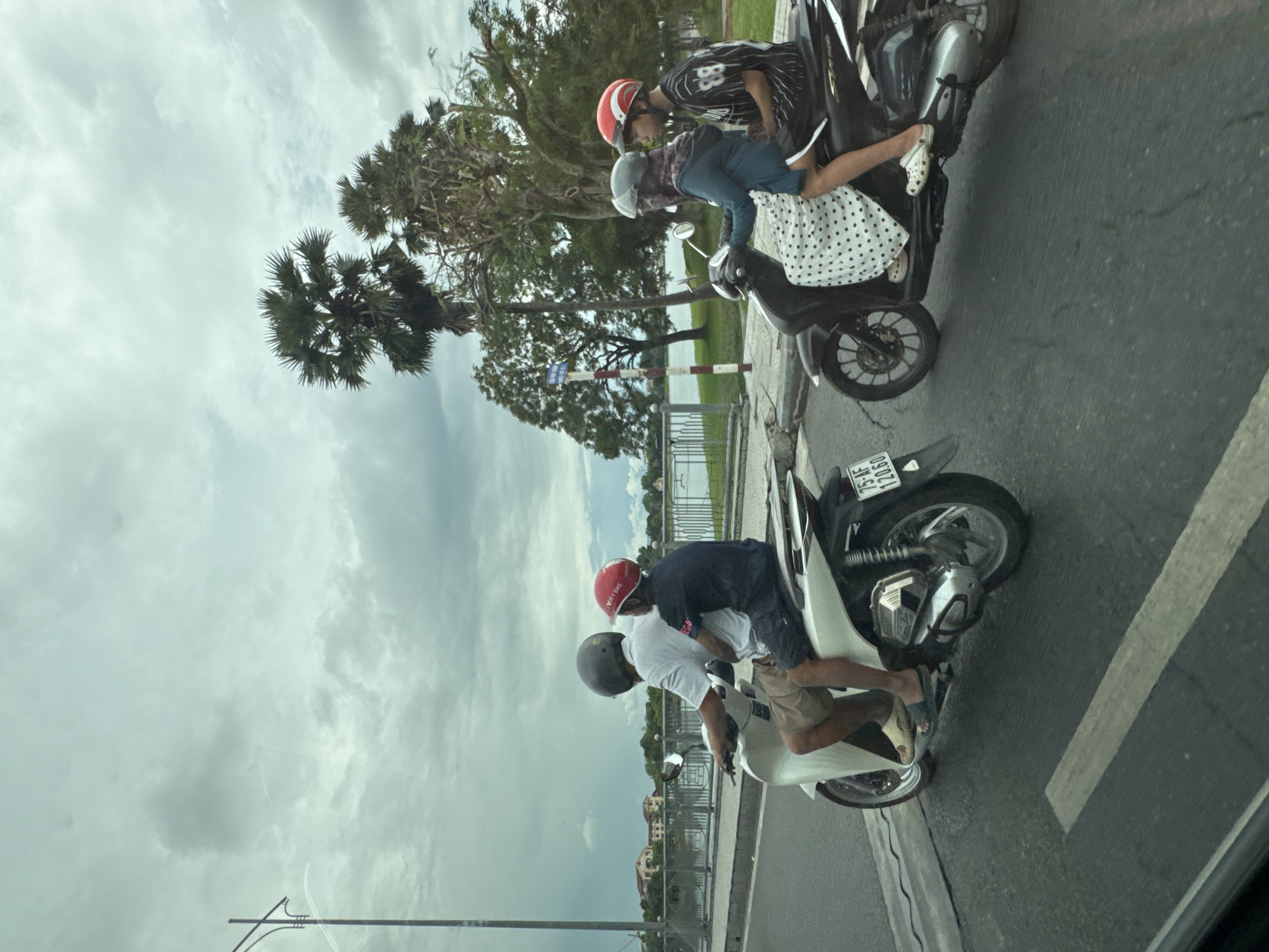 Two motorbikes are traveling on a street in Thuận Hóa Ward, Vietnam. On the left, a white motorbike carries two men. The driver wears a white shirt and khaki shorts, with a dark gray helmet. The passenger wears a dark blue t-shirt, dark shorts, and a red helmet. They are on a street with a grey divider and a pedestrian crossing. In the background, a body of water is visible, with trees and buildings along the shore. On the right, a black motorbike carries two people, likely a woman and a man. The driver, possibly female, wears a blue hooded jacket and a patterned white dress, with a grey helmet and a scarf covering her face. The passenger, possibly male, wears a black and white striped t-shirt and dark shorts, with a red helmet. They are also on the street, with a grassy area and trees beyond. The sky is overcast and cloudy.