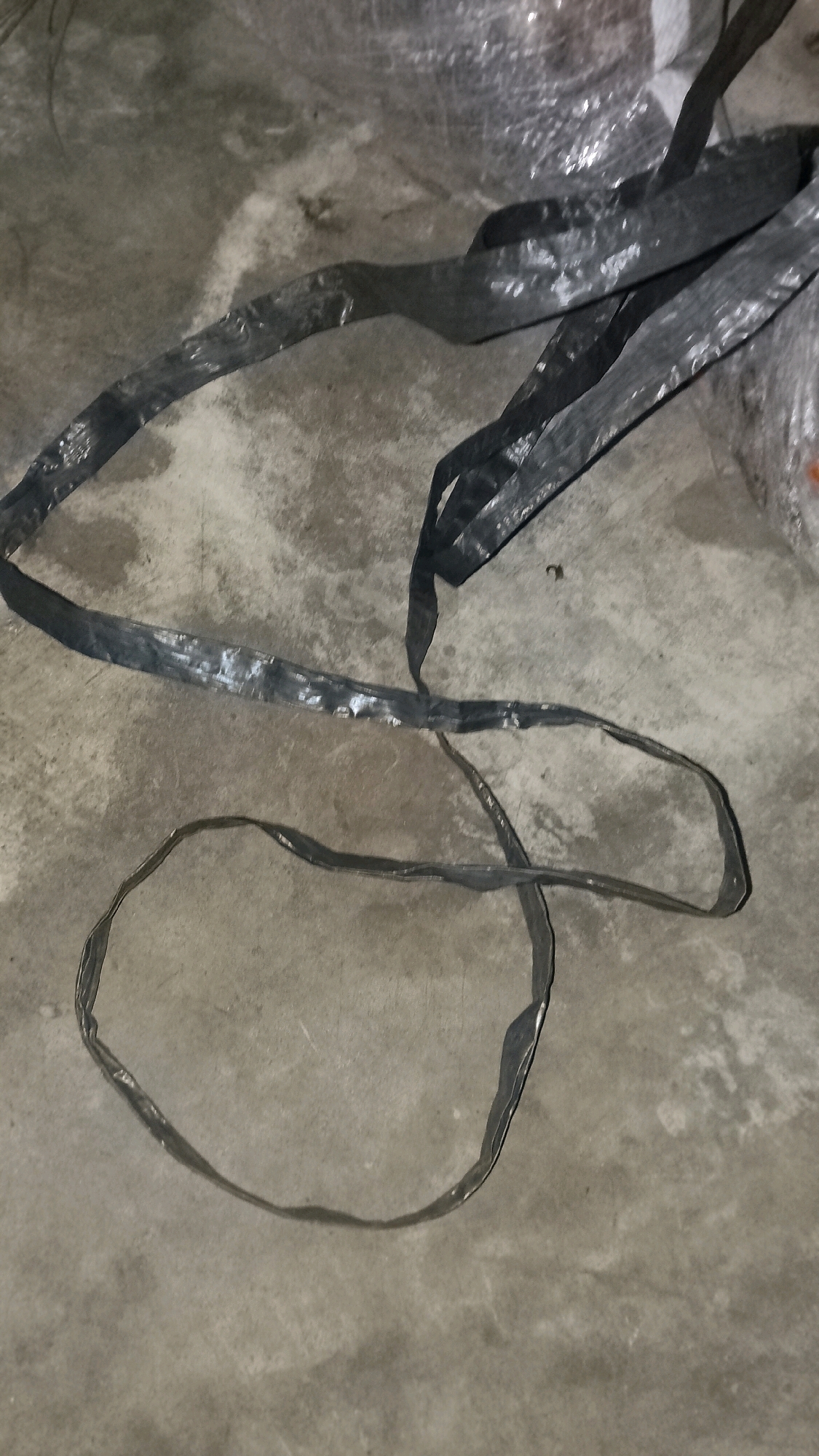 The image shows a black, flat, flexible material, possibly a strap or webbing, lying on a concrete surface. The material is wrinkled and appears to be somewhat worn. It is arranged in a loose coil and a long, meandering line across the ground. In the background, there is a hint of other objects, possibly also wrapped in plastic, which are out of focus. The concrete floor appears to be slightly damp in places. The scene is indoors, possibly a storage area or a workshop. Without further information or context, the specific location within Mulyo Haji, Indonesia, cannot be determined from the image alone.