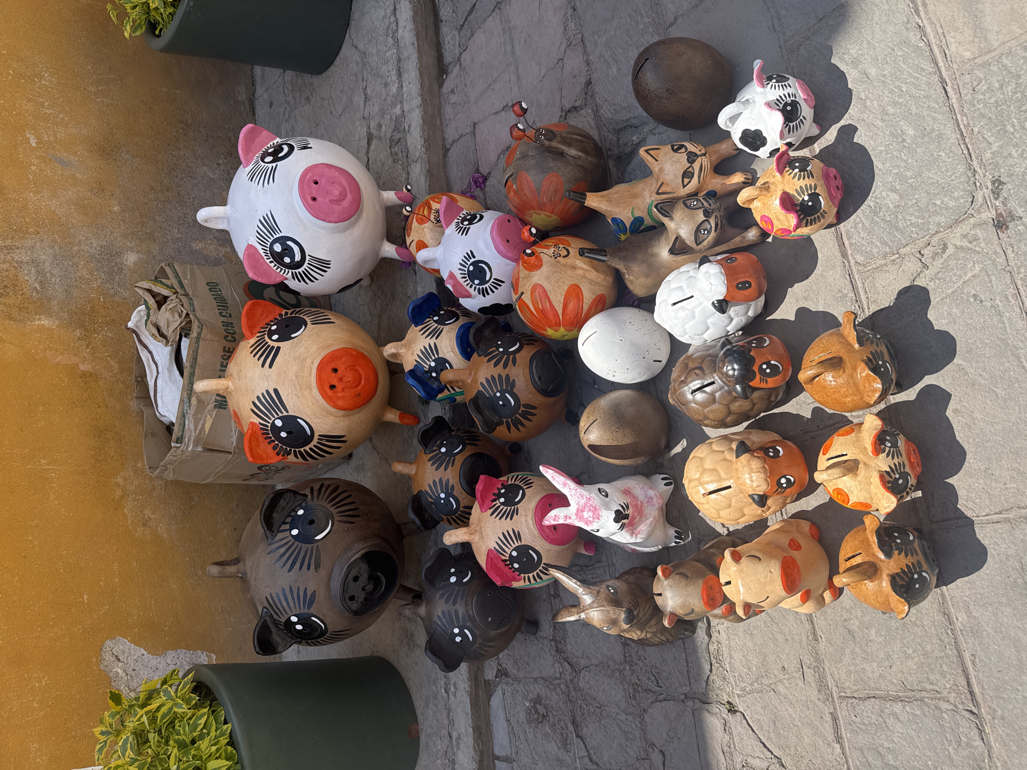 The image depicts a collection of handcrafted ceramic piggy banks and animal figurines arranged on a cobblestone surface. The items vary in color and design, showcasing a range of artistic styles, including pigs, cats, rabbits, owls, and sheep. Some figurines have painted features, while others are left with a natural ceramic finish. They appear to be displayed for sale. The image was taken outdoors, on a sunny day, in Bernal, Mexico. The items are in a random assortment, some stacked on each other. There is a potted plant visible in the lower left corner of the frame.