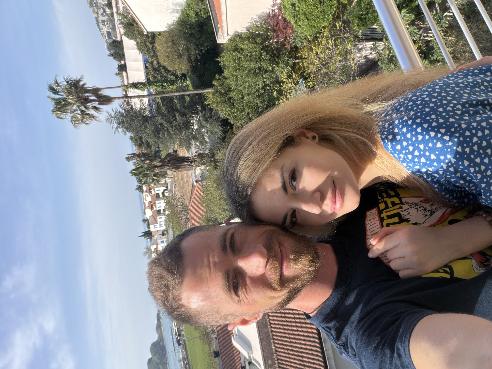 A waist-up shot shows a fair-skinned man and woman. The man has a buzz cut, a beard, and is wearing a dark blue t-shirt. The woman is blond and is wearing a blue dress with small white hearts printed on it. They are standing very close to each other. The man is holding the camera for a selfie. The background of the shot includes a mix of greenery, buildings, and a cloudy blue sky. There are palm trees visible. The location appears to be Budva, Montenegro.