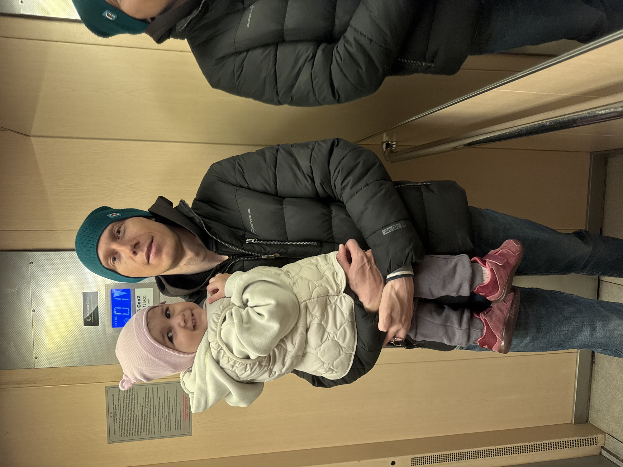 A man holds a smiling baby inside an elevator, likely located in Tobolsk, Russia. The man, with a slight smile, wears a dark puffer jacket with "OUTVENTURE" and "ADD DRY" logos, blue jeans, and a teal knit beanie. The baby, also smiling directly at the camera, is dressed warmly in a light-colored quilted vest, a light pink knit beanie, grey pants, and pink shoes. They are positioned centrally in the elevator, appearing to be taking a photo together.
The elevator interior features light wooden or beige-colored walls and a metal handrail visible on the right. On the left wall, there is an elevator control panel and an information plaque. Text visible on these elements includes "OTIS" (the elevator manufacturer), "ReGen Drive," and "Gen2," along with "13 ПАСС." (indicating a 13-passenger capacity) and "2017". A prominent sign in Russian reads "ПРАВИЛА ПОЛЬЗОВАНИЯ ПАССАЖИРСКИМИ ЛИФТАМИ," which translates to "RULES FOR USING PASSENGER ELEVATORS," confirming the Russian context. The warm attire of both subjects suggests a cool climate. The overall scene conveys a tender and happy moment between the man and the baby.