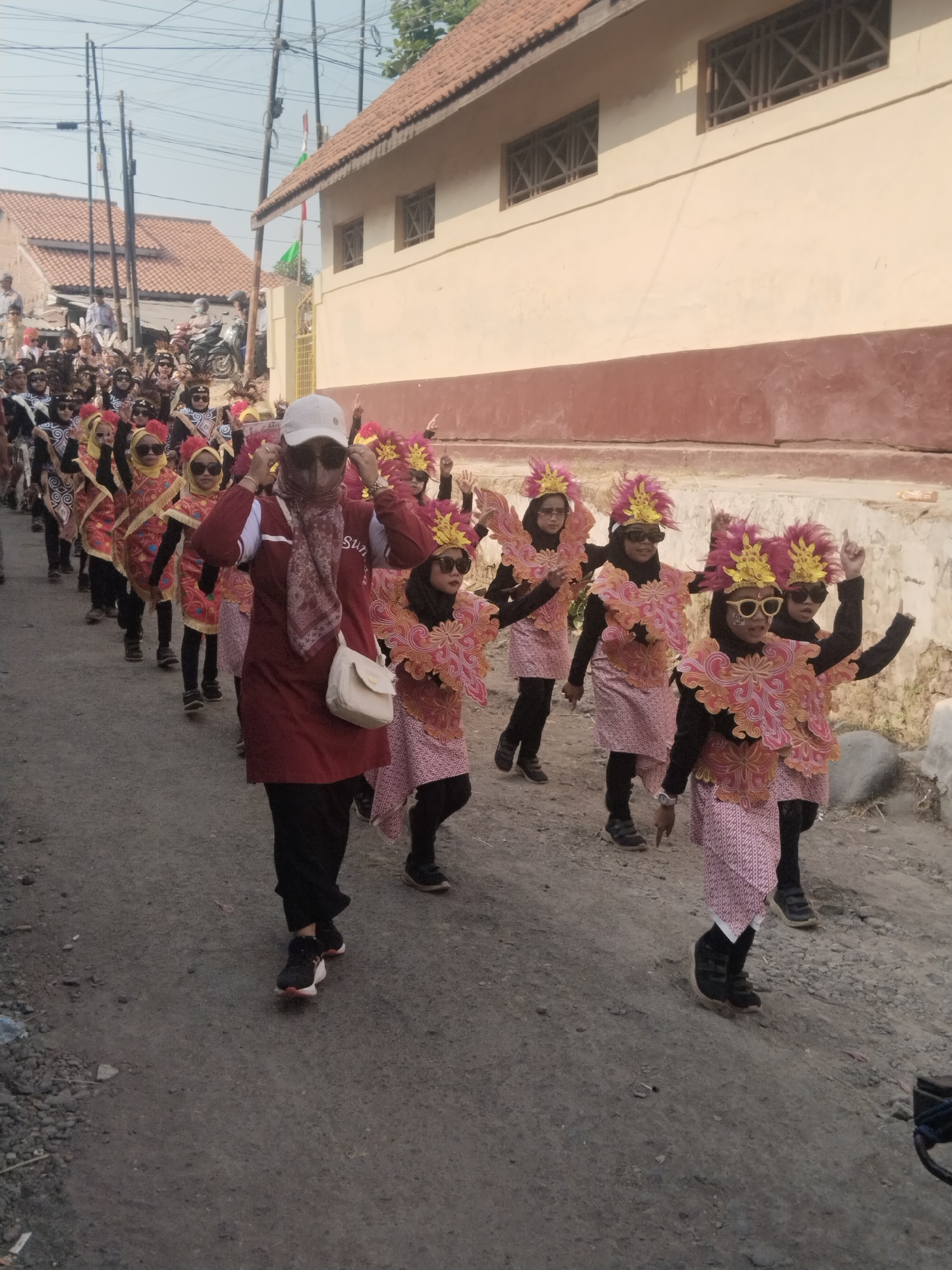 In Jembayat, Indonesia, a group of children wearing vibrant costumes participates in a parade. The costumes consist of pink and orange layered fabric adorned with intricate patterns, black long sleeves and pants, and a headdress featuring pink feathers and yellow accents. Most of the children also wear sunglasses. Leading the group is an adult woman in a maroon top, black pants, a white cap, sunglasses, and a patterned scarf. She carries a white purse on her shoulder and is adjusting her cap and sunglasses. The parade proceeds down a dirt road lined with buildings. The buildings have tiled roofs and cream-colored walls with dark brown trim. Utility poles and wires are visible overhead. The ground is rough and covered in loose gravel and debris. The overall scene suggests a festive cultural event taking place in a semi-urban or rural setting.