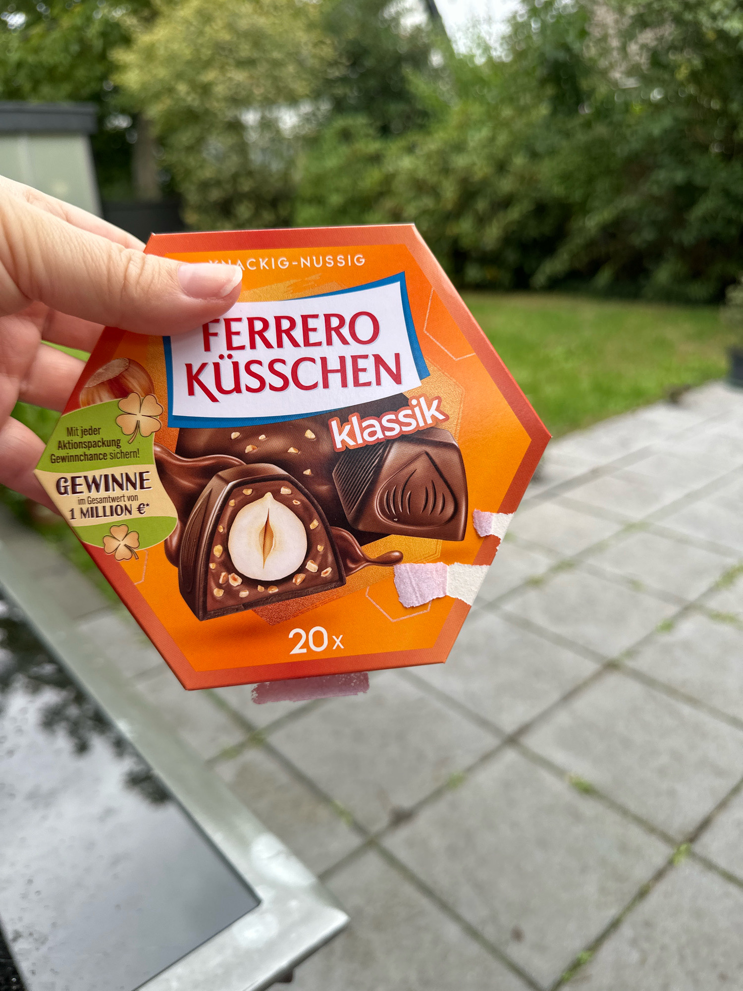 The image shows a hand holding a hexagonal orange box of Ferrero Küsschen Classic chocolates outdoors. The box has a picture of the chocolates and text that includes "Ferrero Küsschen", "knackig-nussig" (crunchy-nutty), "klassik", and a promotion that says "GEWINNE im Gesamtwert von 1 MILLION €*" (Win a total value of 1 million €*). The background consists of greenery, a patio with square pavers, and a glimpse of a building. The image appears to be taken in a residential or garden setting, possibly Krefeld, Germany, given the German text on the box.