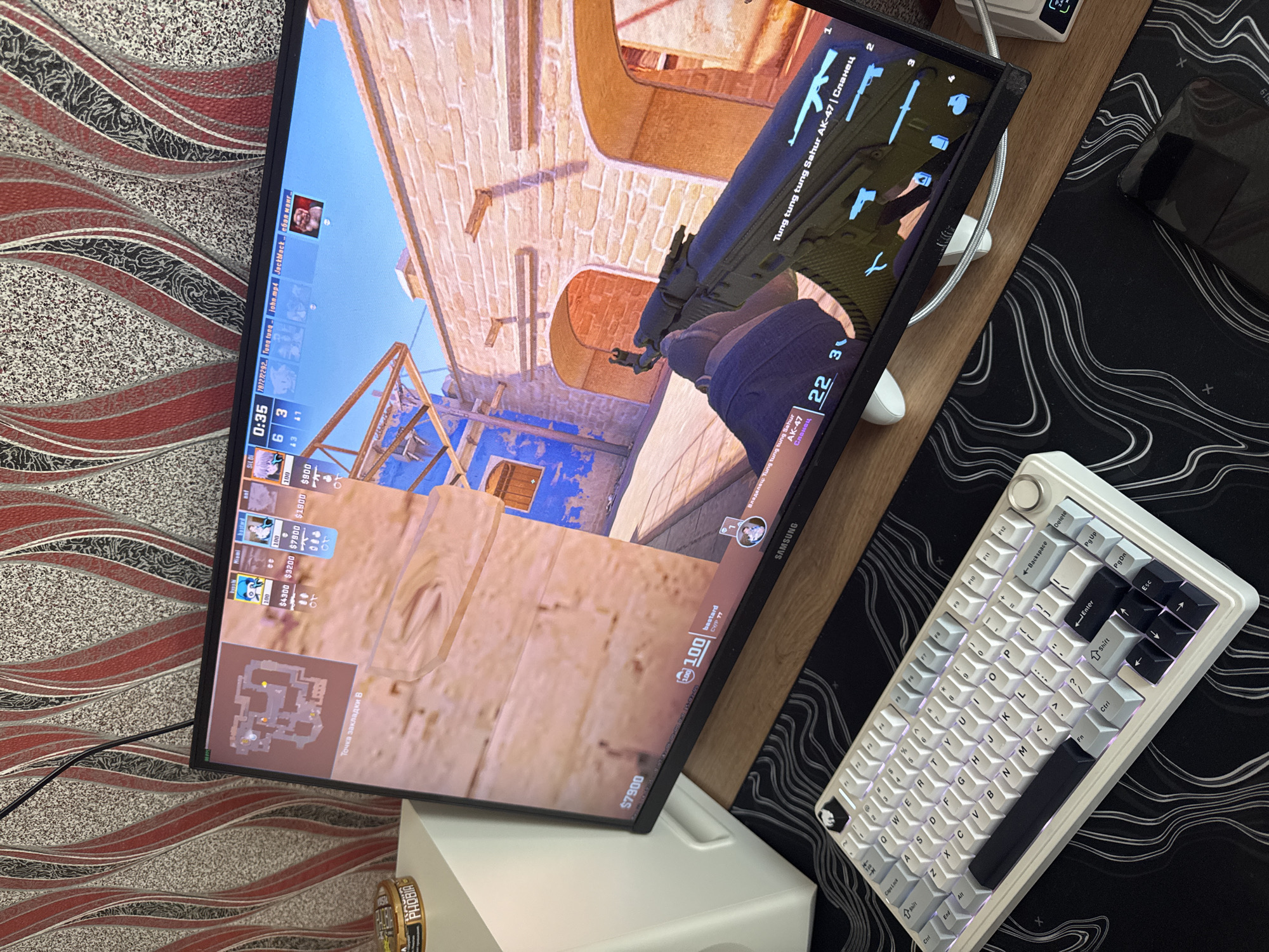 A monitor displays gameplay of Counter-Strike: Global Offensive, showing a first-person view of a player holding an AK-47 in what appears to be the "Dust II" map. In-game elements include a score tracker of 6 to 3, timer at 0:35, player names, money amounts. Below the monitor stands a white box, and on the desk is a white mechanical keyboard with gray and black keycaps on a black and white patterned desk mat. The setting is most likely indoors on a desk, with a textured background on the left.