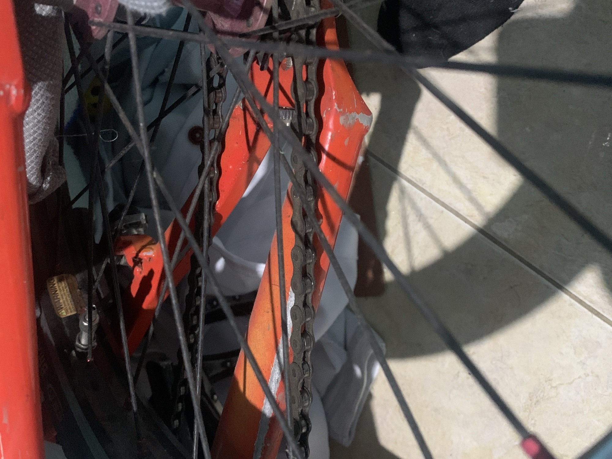 A close-up, tilted shot captures a segment of a worn bicycle, viewed obliquely through its wheel spokes. The primary subject is a vibrant orange-red bicycle frame, exhibiting noticeable wear and tear with chipped paint, particularly on the chainstay. A metallic, somewhat grimy bicycle chain runs parallel to the frame, partially obscured by the numerous thin, dark spokes that crisscross the foreground, forming a dense pattern.
Behind the spokes and chain, glimpses of various elements can be seen. A silver valve stem is clearly visible on the wheel, with a small yellow sticker nearby that bears the word "WARNING". Further back, white and grey fabric or cloth is bundled or draped, and indistinct darker shapes suggest other background objects.
The scene is bathed in bright sunlight, casting sharp, elongated shadows from the spokes and bike frame onto a light-colored tiled floor. This strong illumination indicates it is daytime, likely midday or early afternoon, outdoors or in a space receiving direct sun. No specific activity is depicted; the bicycle appears stationary, possibly parked or stored. The overall impression is one of a well-used bicycle in a casual, sunlit environment.