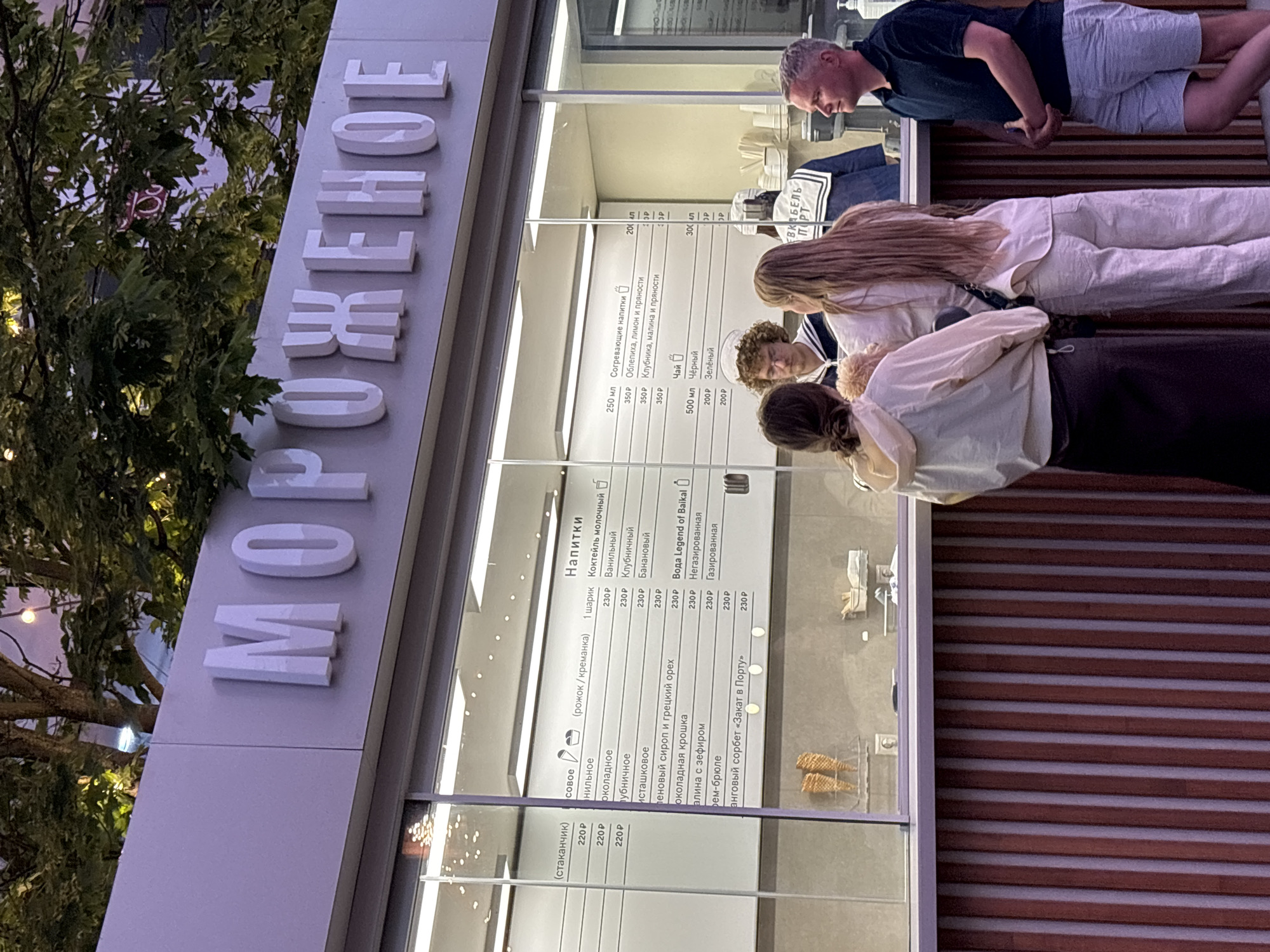 The image shows an outdoor ice cream stand in Saint Petersburg, Russia, with the word "МОРОЖЕНОЕ" (ice cream) prominently displayed on a sign above the counter. The stand features a glass display window showcasing a menu of ice cream flavors and drinks, along with prices. Several people are visible at the counter, appearing to order or wait for their items. One person in a sailor-style uniform is behind the counter, likely an employee. The overall environment suggests a warm evening, with some decorative lighting visible. The sign's Cyrillic script and the architectural style are consistent with locations in Russia.