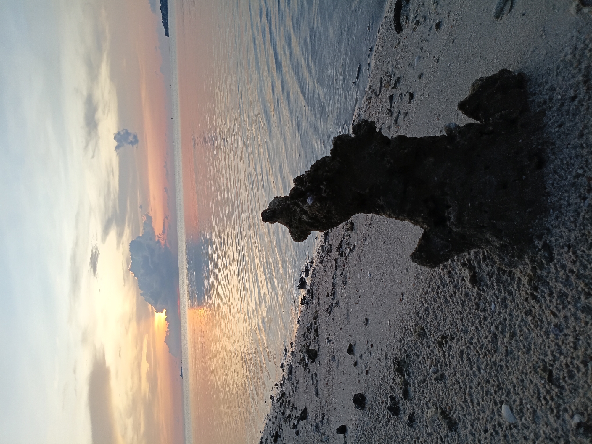 The image depicts a beach scene in Baan Wok Tum, Thailand, during a sunset. The foreground consists of a sandy beach with scattered rocks and debris. A large, dark rock formation stands prominently on the shoreline, partially obscuring the view. Beyond the beach, the water reflects the colors of the sunset. The sky displays a gradient of colors, ranging from light blue to orange and pink, with clouds partially obscuring the sun. The overall scene suggests a tranquil and natural environment.