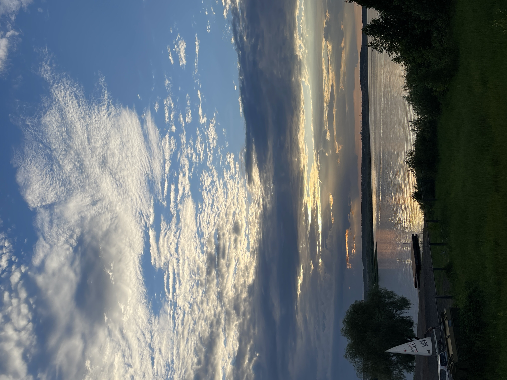 The image depicts a serene waterscape at sunset, with a wide expanse of water reflecting the warm hues of the sky. The sky is a canvas of dramatic clouds, ranging from wispy cirrus formations to darker, more substantial cloud banks, all bathed in the golden light of the setting sun. Rays of sunlight pierce through the clouds, creating a luminous effect on the water's surface.
On the right side of the frame, a lush green embankment slopes down towards the water, with dark green trees providing a natural border. The water itself appears calm, with gentle ripples disturbing its surface. A small wooden dock or pier extends slightly into the water, and a single, small sailboat with a white sail is moored nearby. A small, dark green tree with a weeping canopy stands closer to the water's edge. A portion of a silver car is visible in the lower right corner, and a small white sign with some text is attached to the side of the sailboat. The sign is partially obscured but appears to have "2105" and "RUS" visible.
The overall mood of the image is peaceful and contemplative, evoking the tranquility of nature at dusk. The interplay of light and shadow, the vastness of the sky and water, and the subtle details of the sailboat and its surroundings contribute to a feeling of quiet beauty. The time of day is clearly sunset, given the golden light and the long shadows that would be cast. The weather appears clear with dramatic cloud formations.