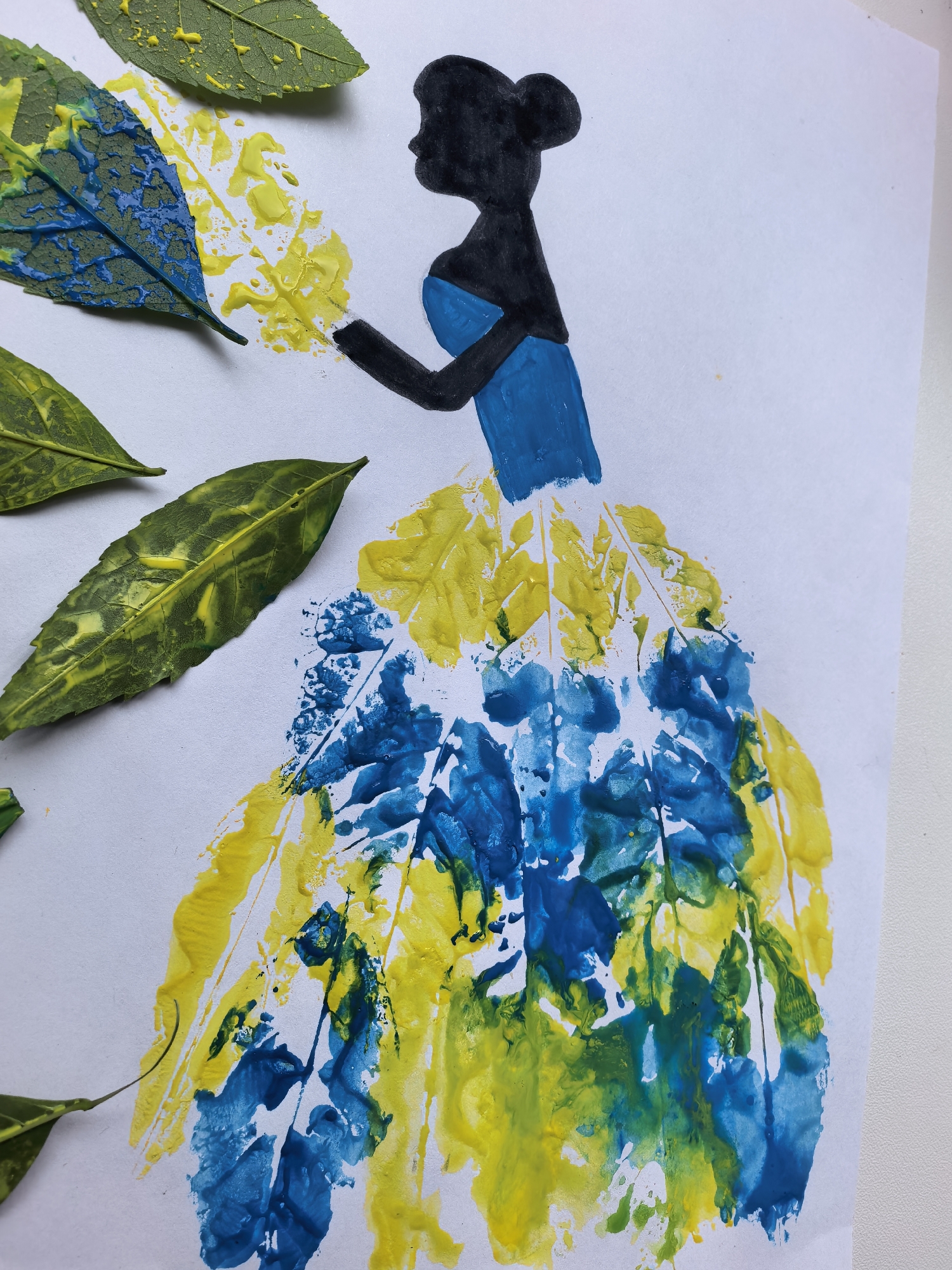 The image depicts a piece of art created using leaves and paint. A black silhouette of a woman, dressed in a blue bodice and a voluminous skirt made of leaf prints in yellow and blue, is the central subject. Several green leaves, some with yellow paint smudges, are arranged around the silhouette, with one leaf in the upper left corner displaying a prominent blue and yellow pattern. The artwork is set against a plain white background. There is no indication of a location in Sterlitamak, Russia, or any other specific geographical context. The scene is a still image of an artistic creation.